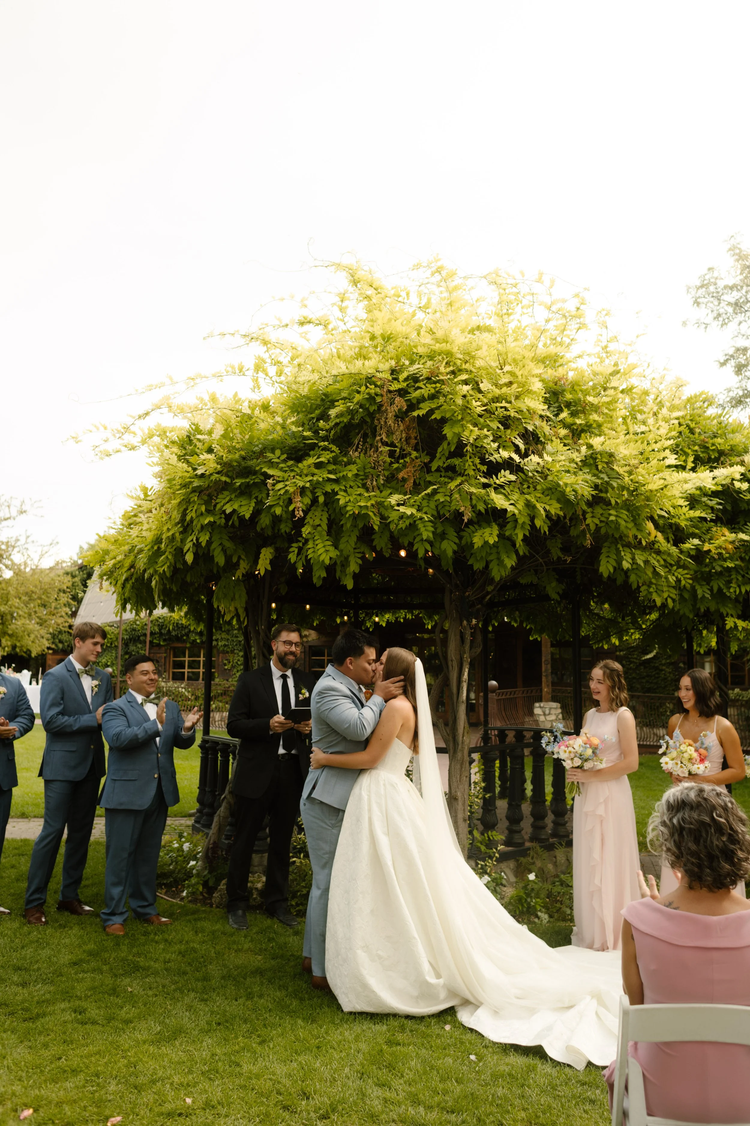 Bride and groom kissing under a lush green gazebo while guests stand and watch during an outdoor ceremony at a romantic utah wedding venue.