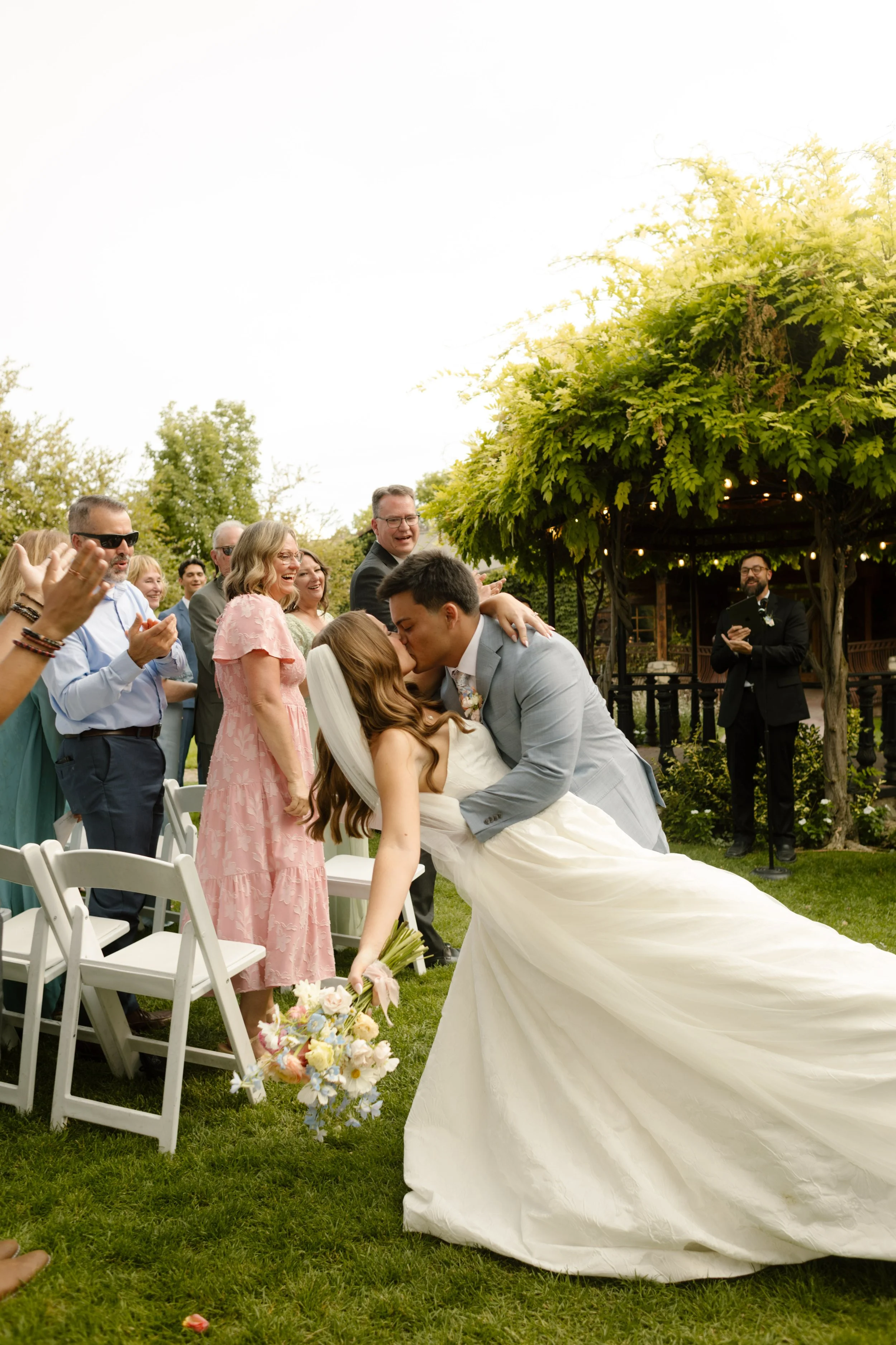 Groom dipping the bride for a kiss in the aisle as guests cheer around them in an outdoor garden setting.