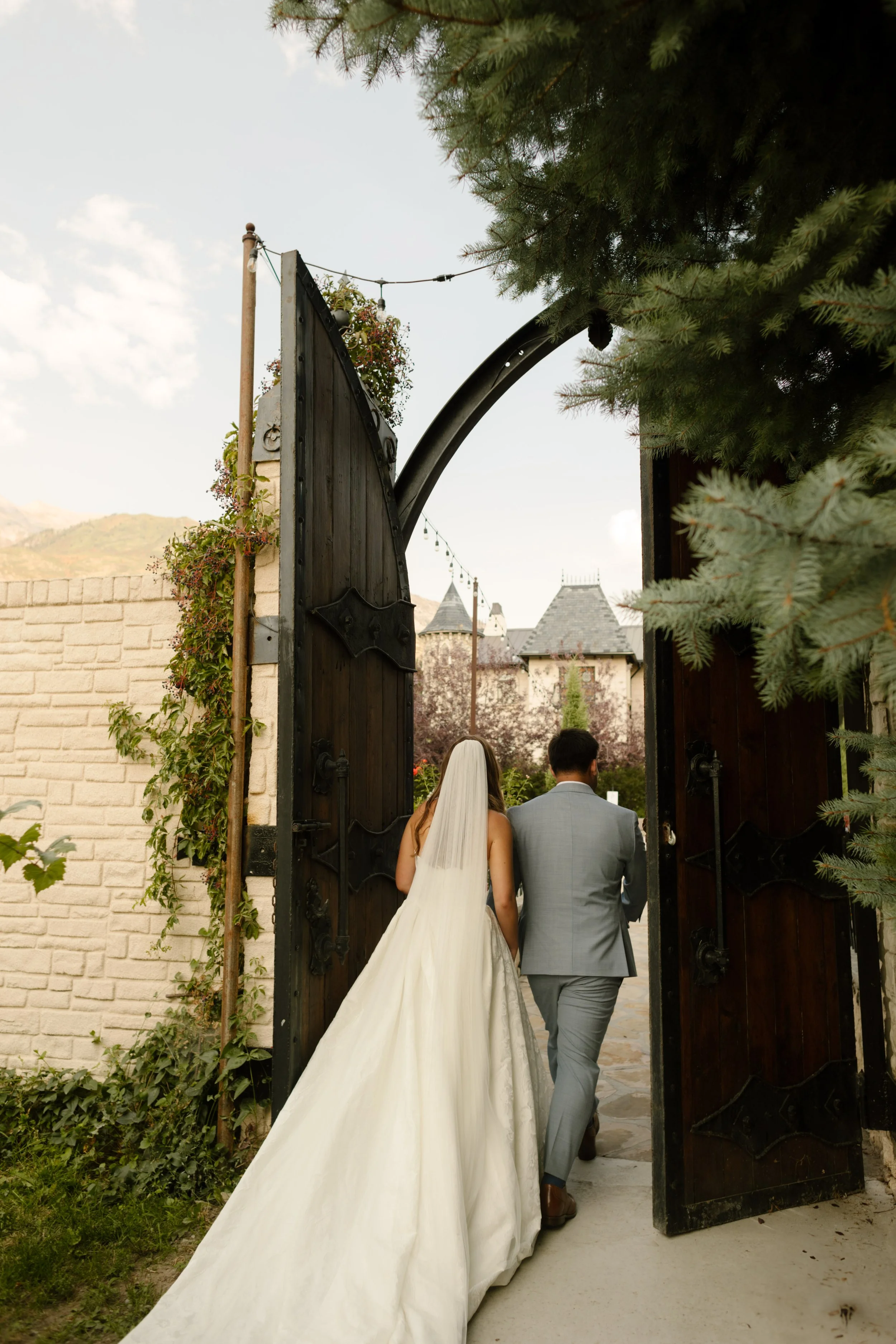 Bride and groom walking through large wooden gates into a courtyard with stone walls and mountain views at a charming utah wedding venue.