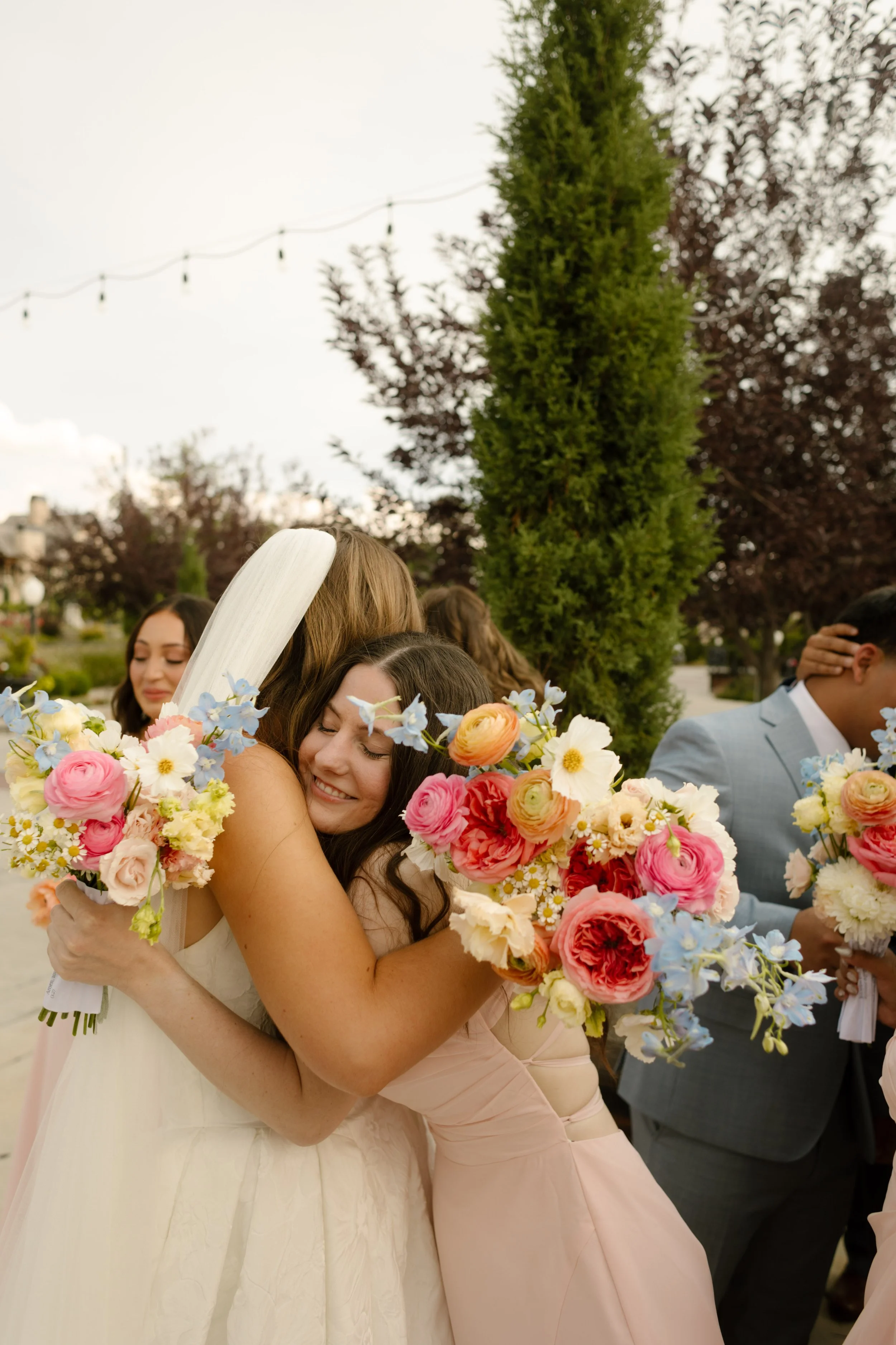 Bride hugging a bridesmaid holding a colorful bouquet of pink, peach, and blue flowers during the ceremony at a garden utah wedding venue.