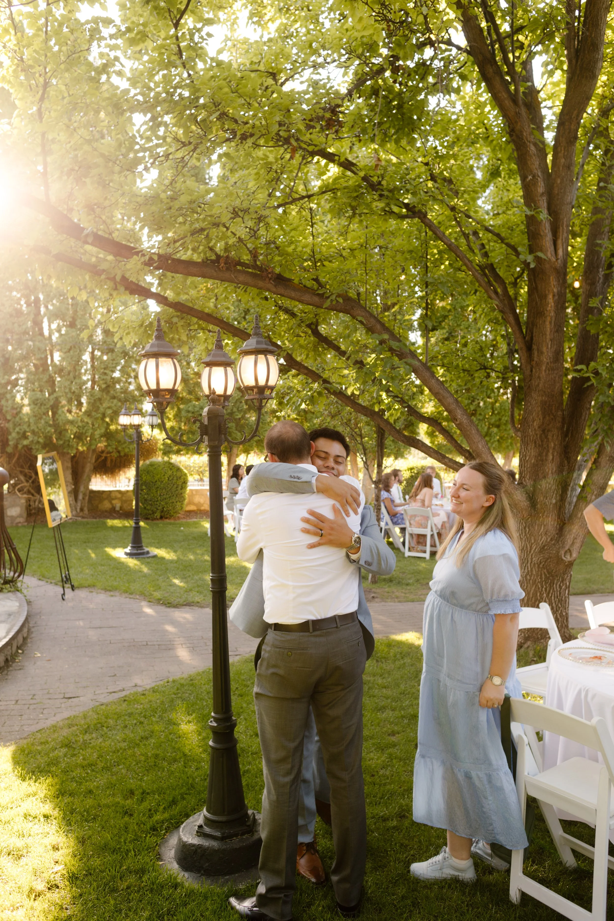 Groom embracing an older guest beneath tall trees and vintage-style lampposts during cocktail hour at a scenic utah wedding venue.
