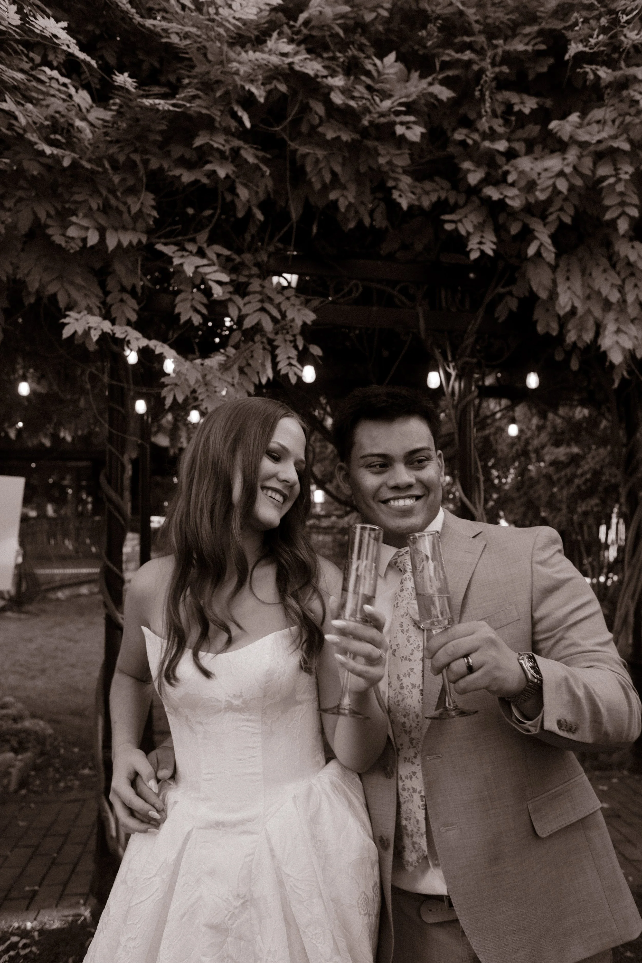 Bride and groom smiling under string lights and trees, holding champagne glasses during golden hour at a romantic utah wedding venue.