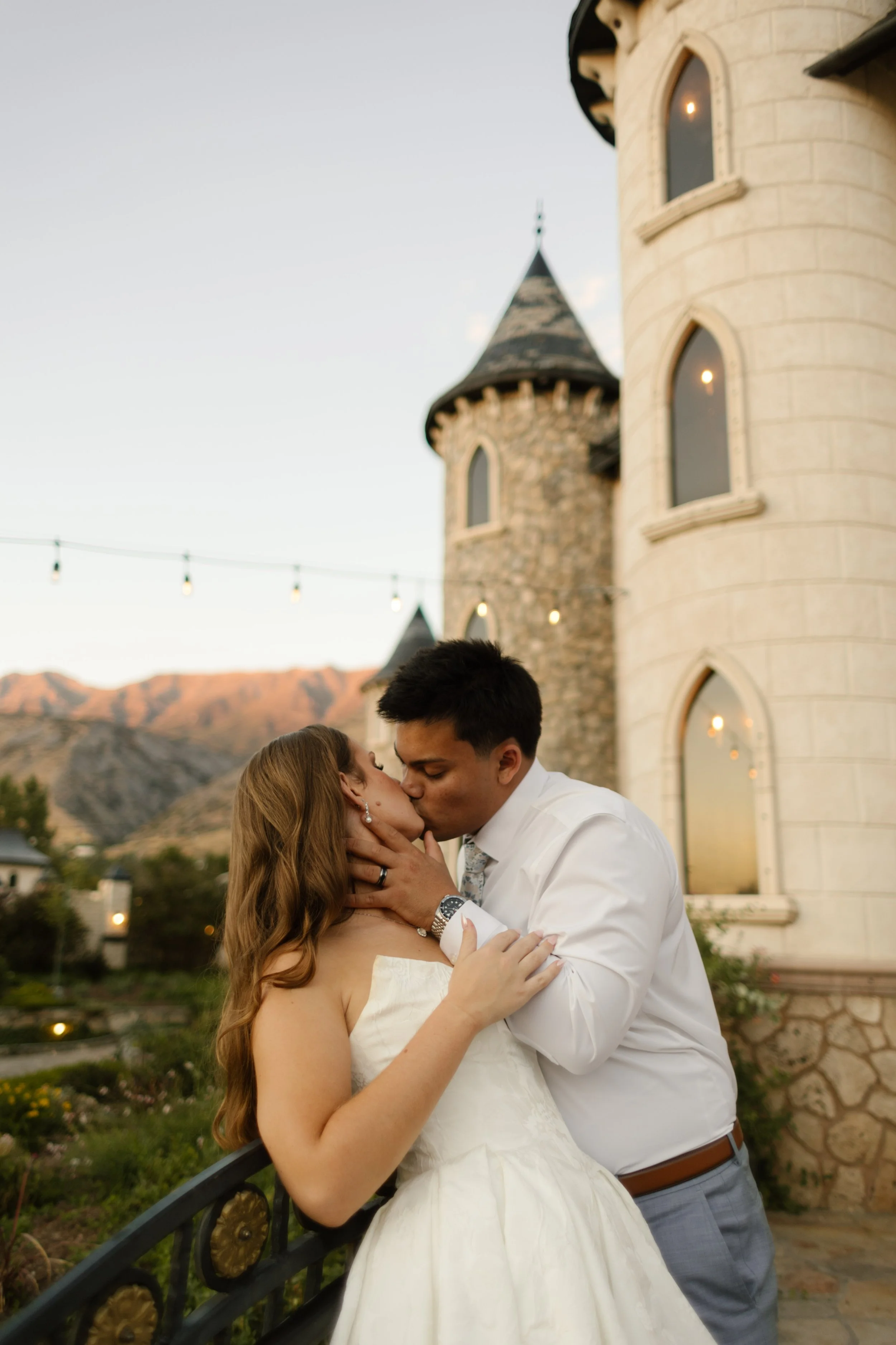 Bride and groom kissing beside a stone castle tower with sunset light hitting the mountains at a dreamy utah wedding venue.