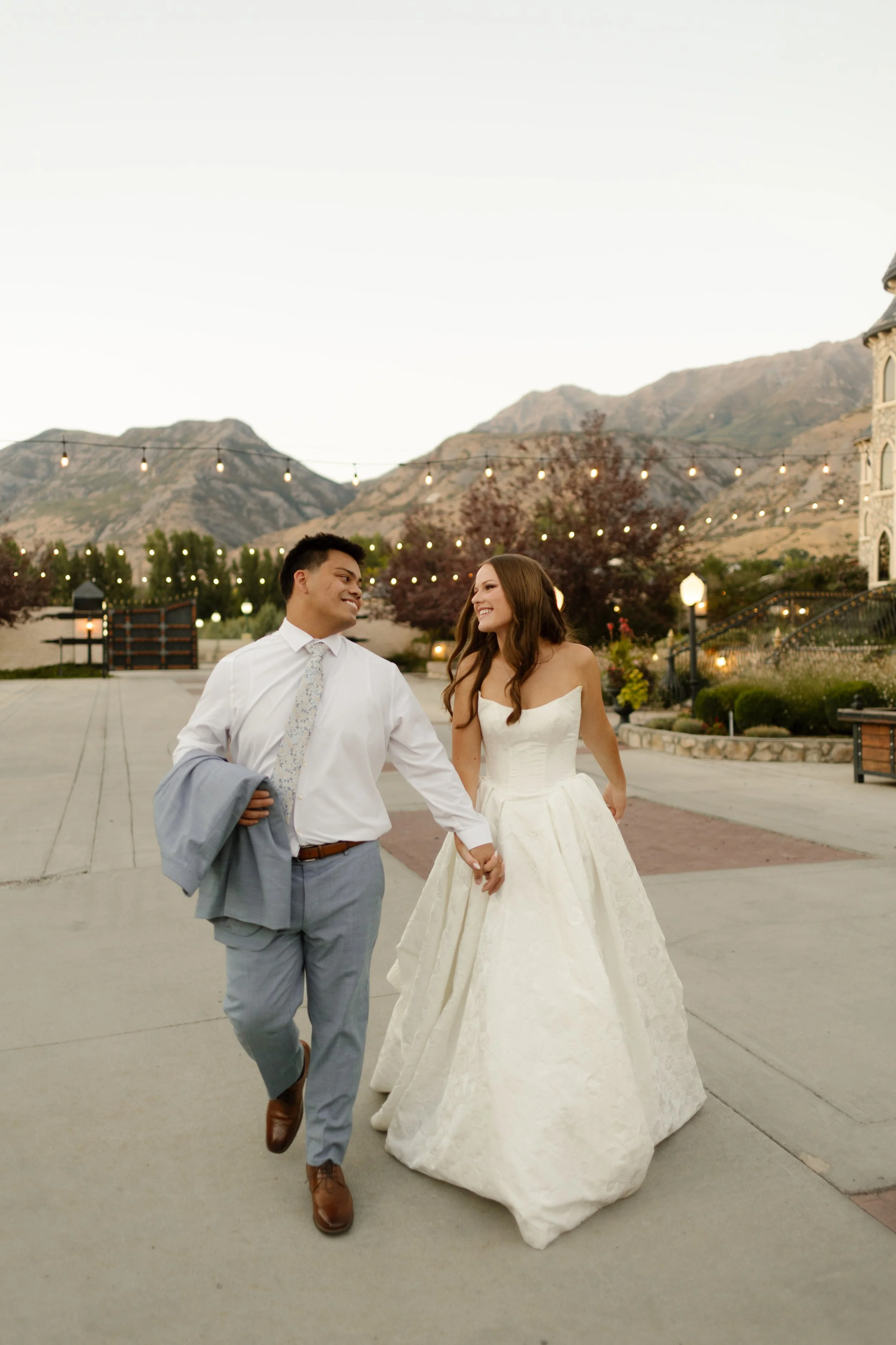 Bride and groom holding hands and laughing with mountains and market lights behind them at a picturesque utah wedding venue.