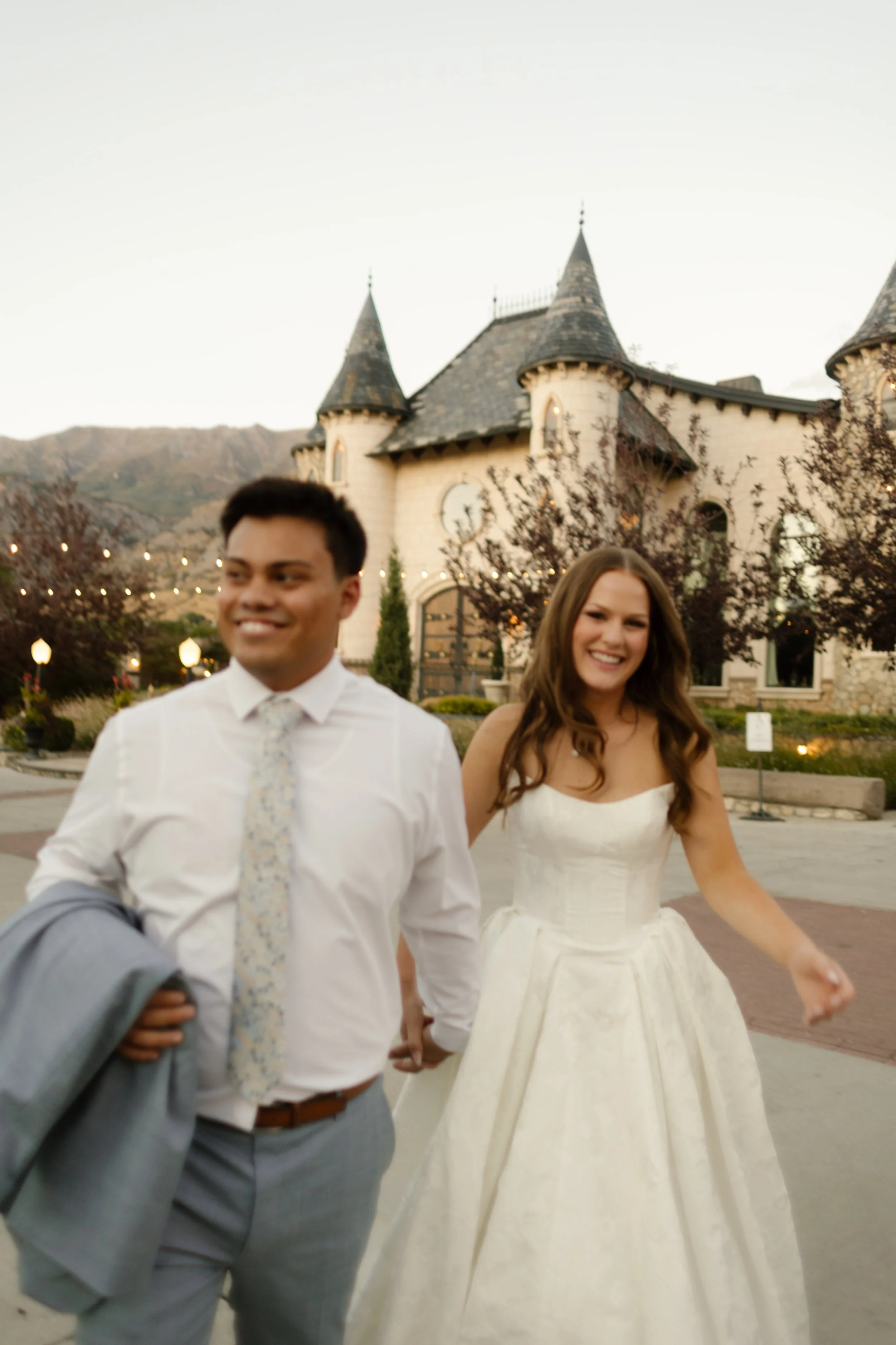Newlyweds walking hand in hand in front of a castle-style building with mountain views at a scenic utah wedding venue.