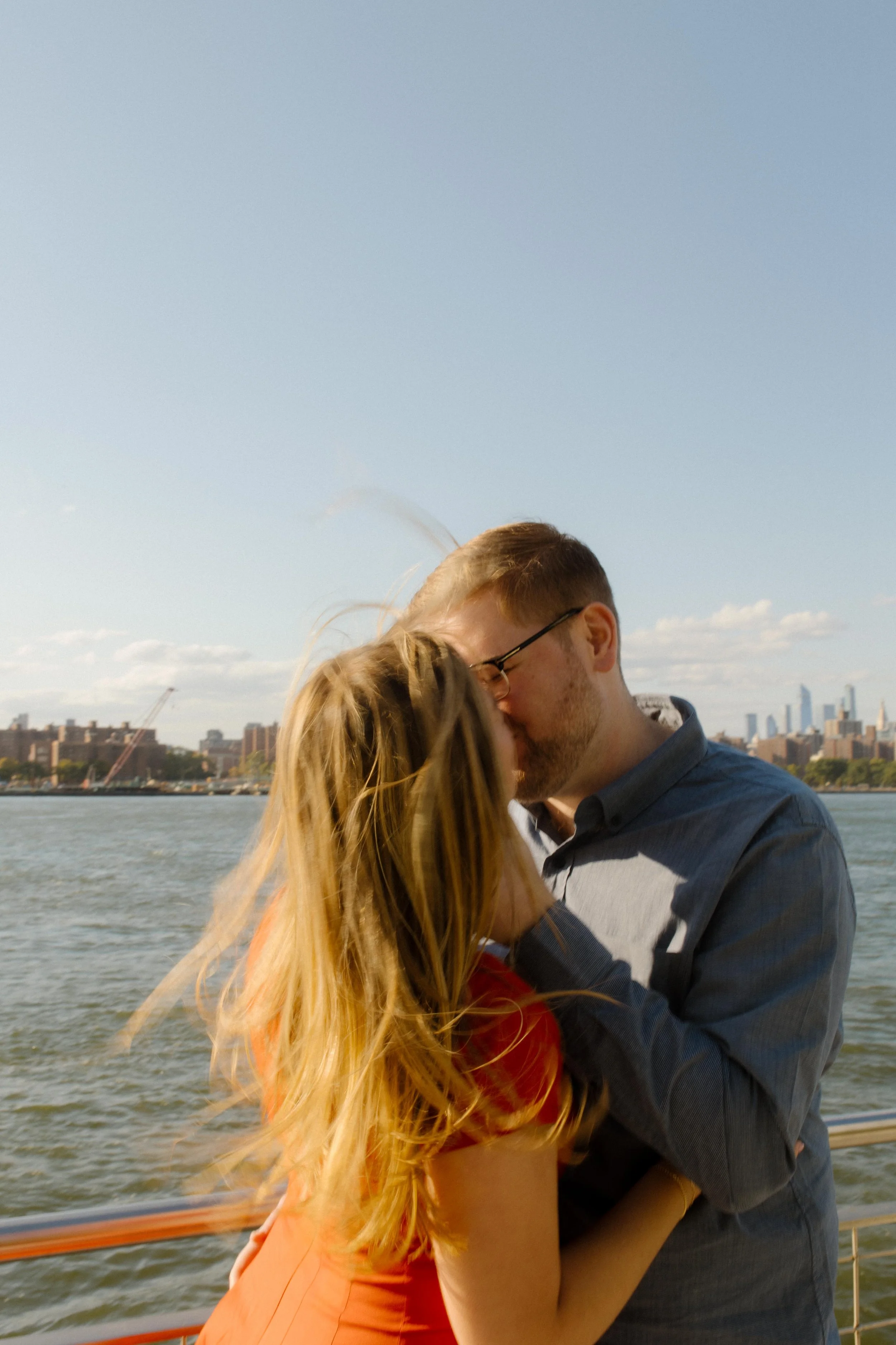 Romantic nyc engagement photos of couple kissing along the waterfront with Manhattan skyline in the distance.