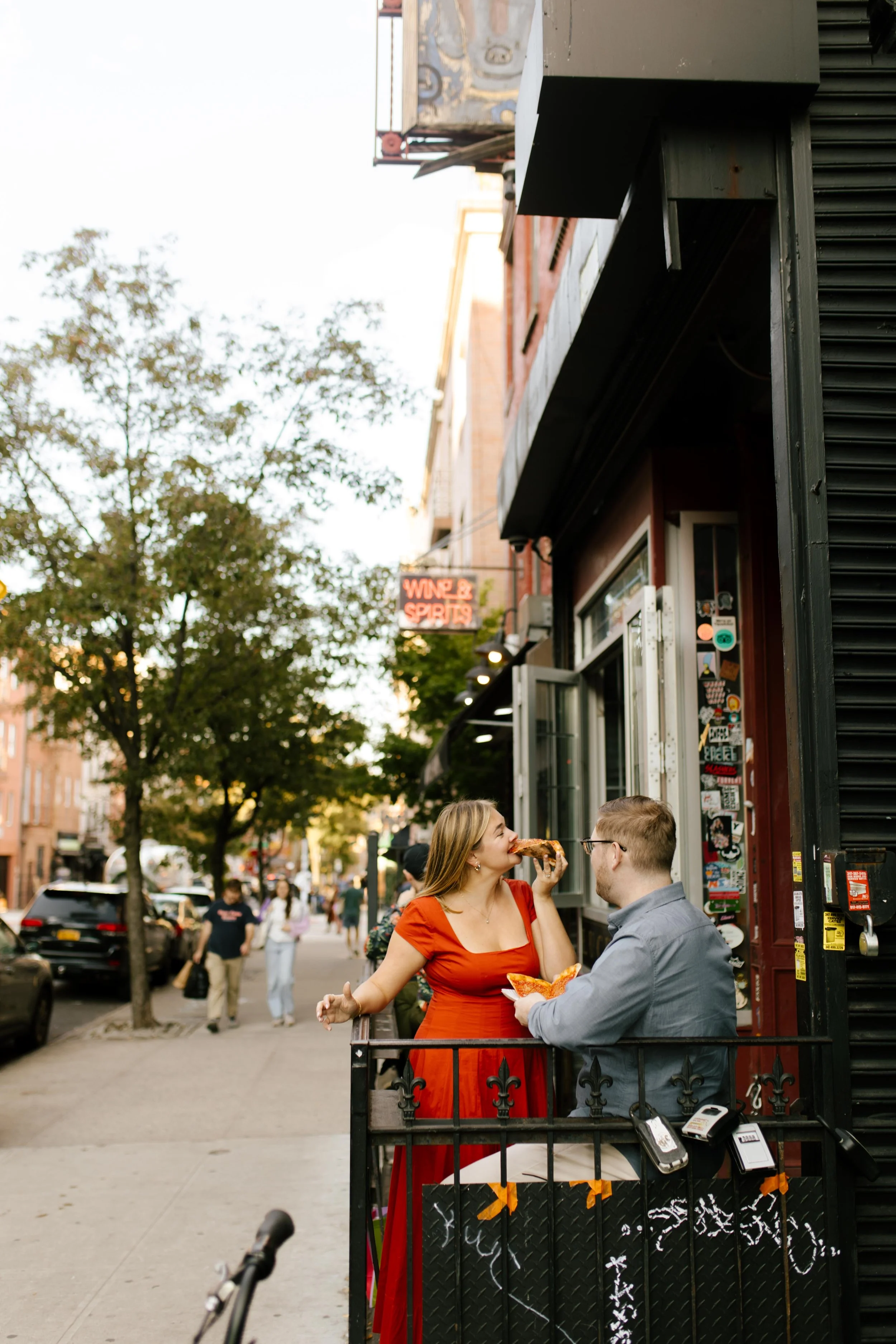 Playful nyc engagement photos outside a Brooklyn pizza spot, bride-to-be in red dress mid-bite on the sidewalk.