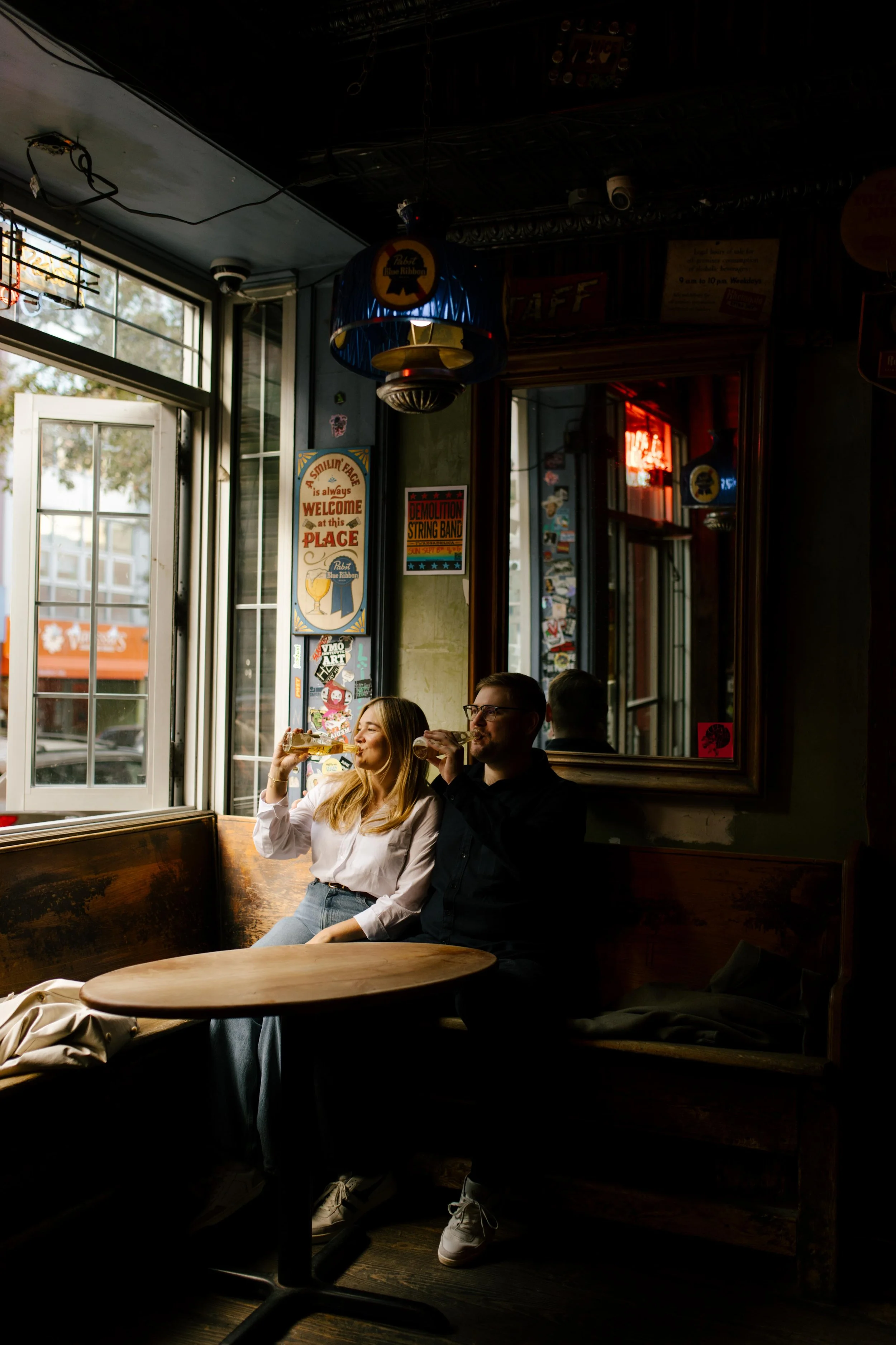 Couple sipping drinks in a cozy Brooklyn bar booth, window light pouring in during their engagement session.