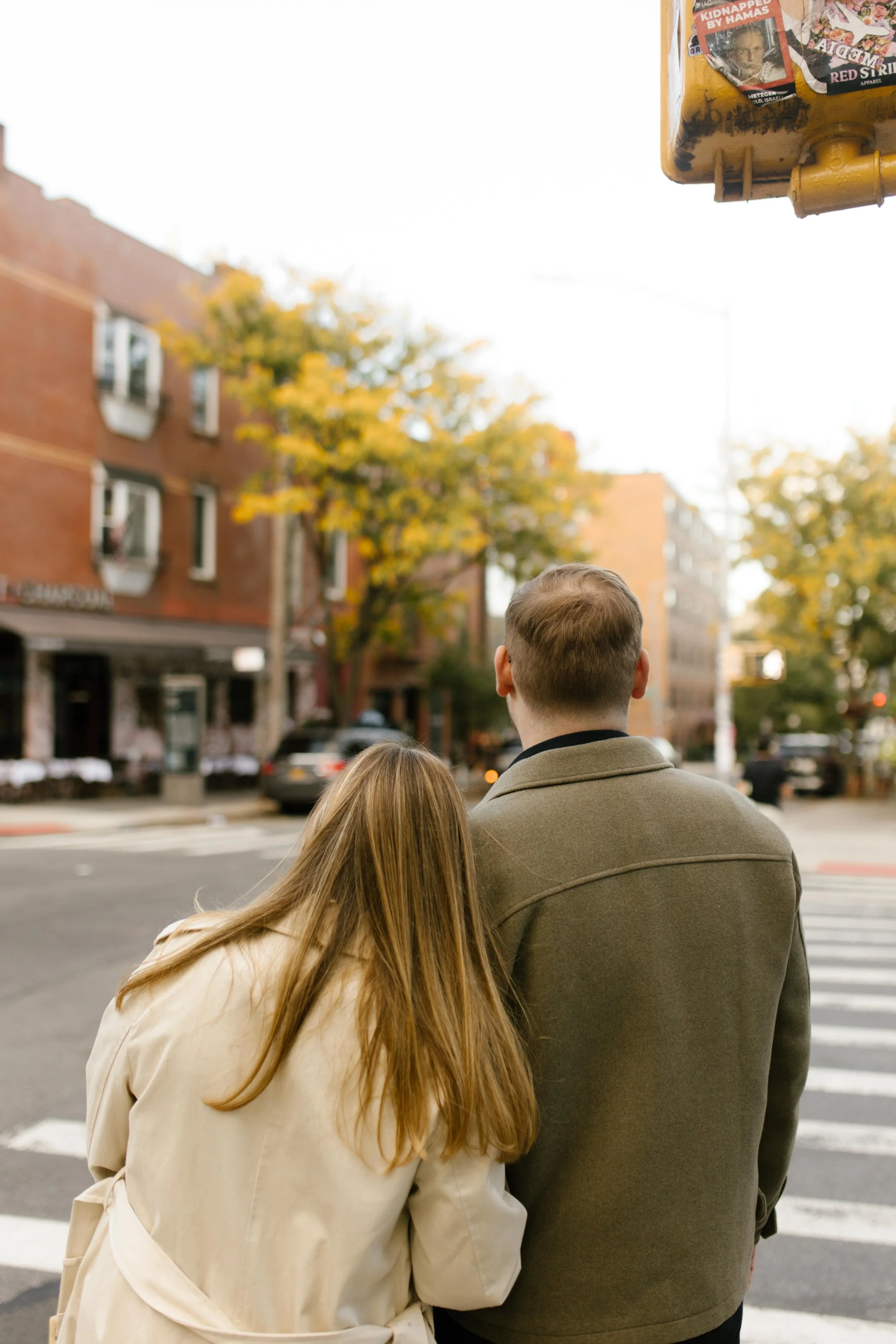 Couple crossing a Brooklyn street in fall, leaning into each other beneath golden trees and brick buildings.