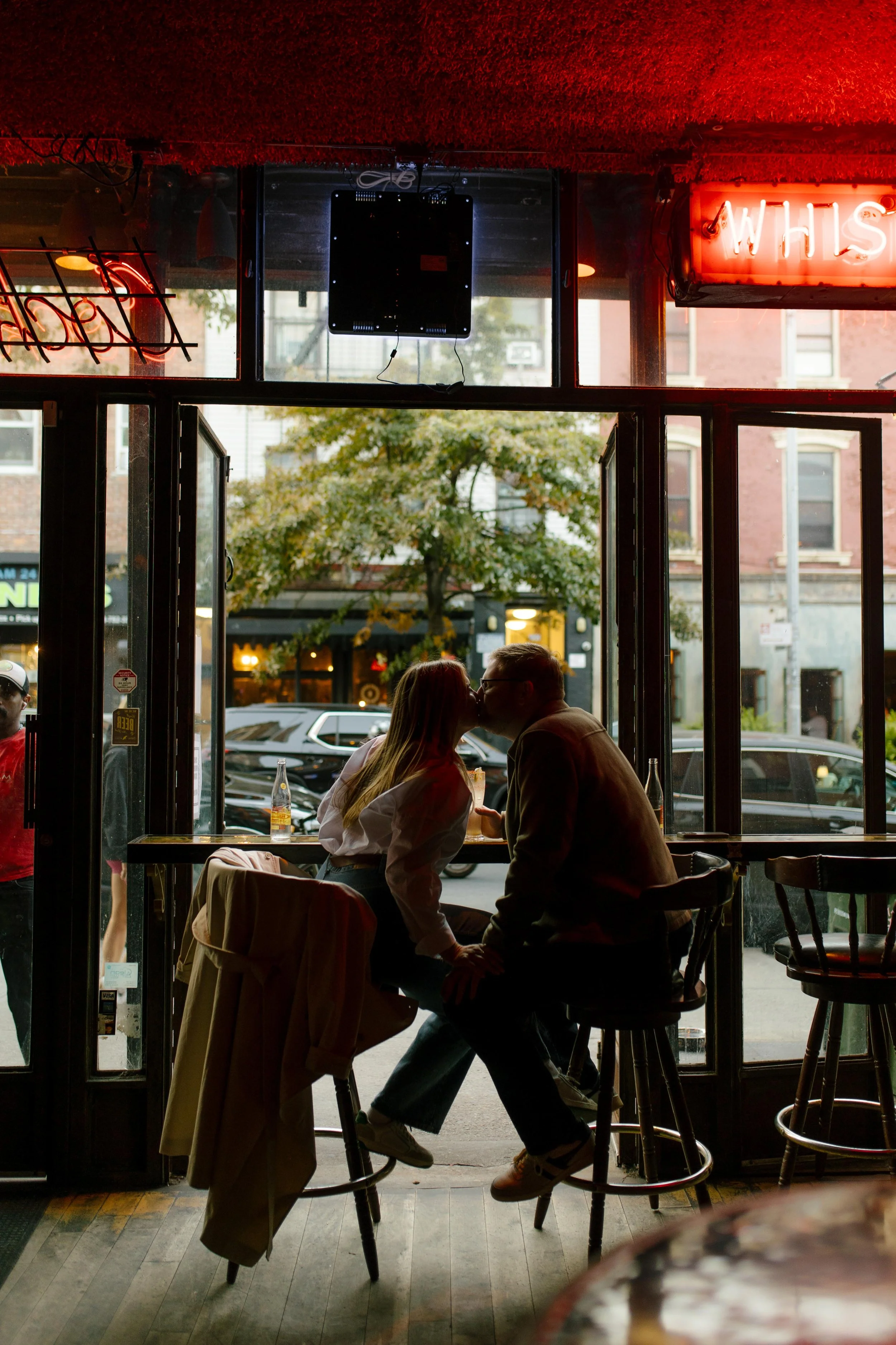 Moody nyc engagement photos of couple kissing at a window bar seat under glowing red neon lights.