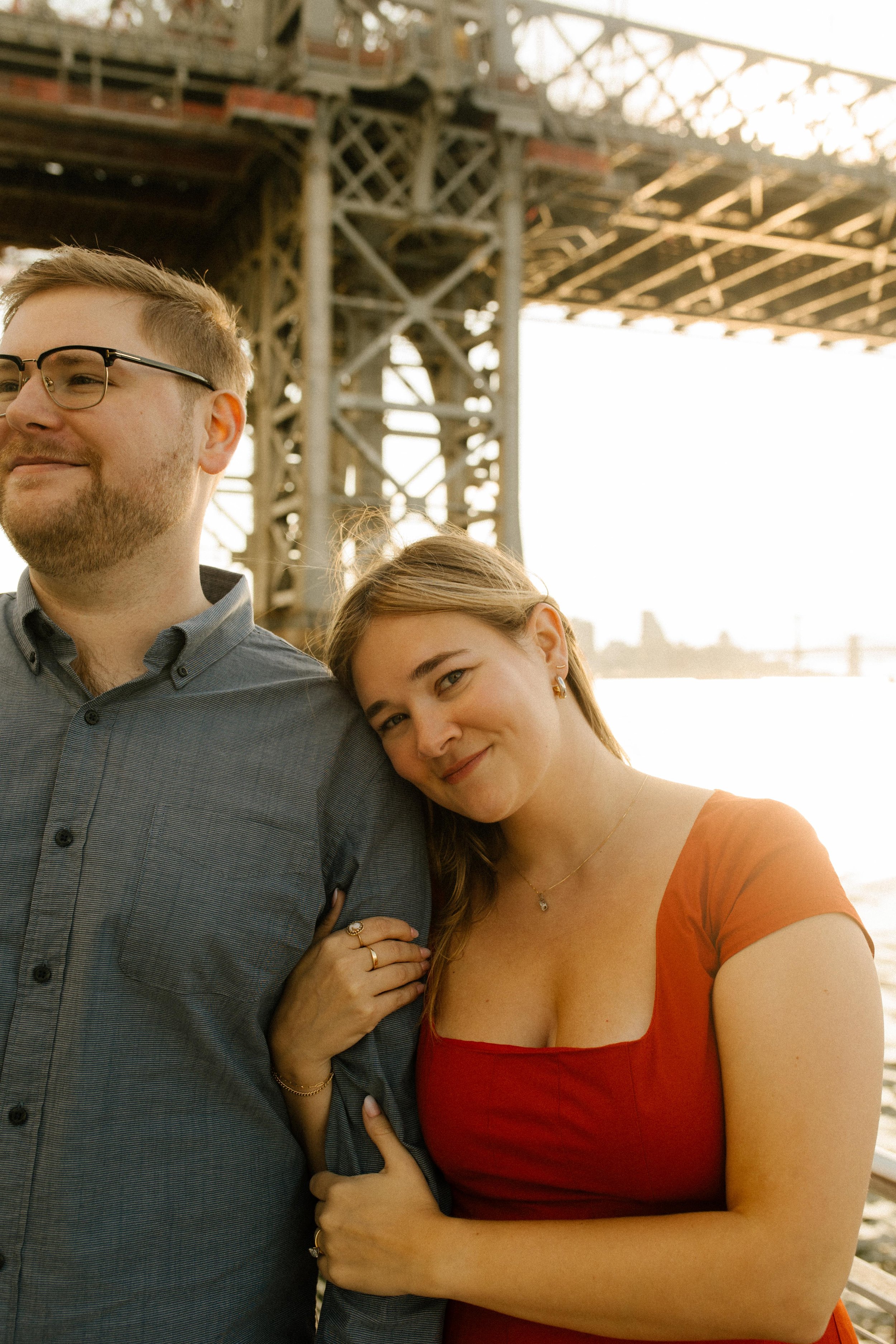 Romantic portrait under the Manhattan Bridge at sunset, her red dress glowing in soft evening light.