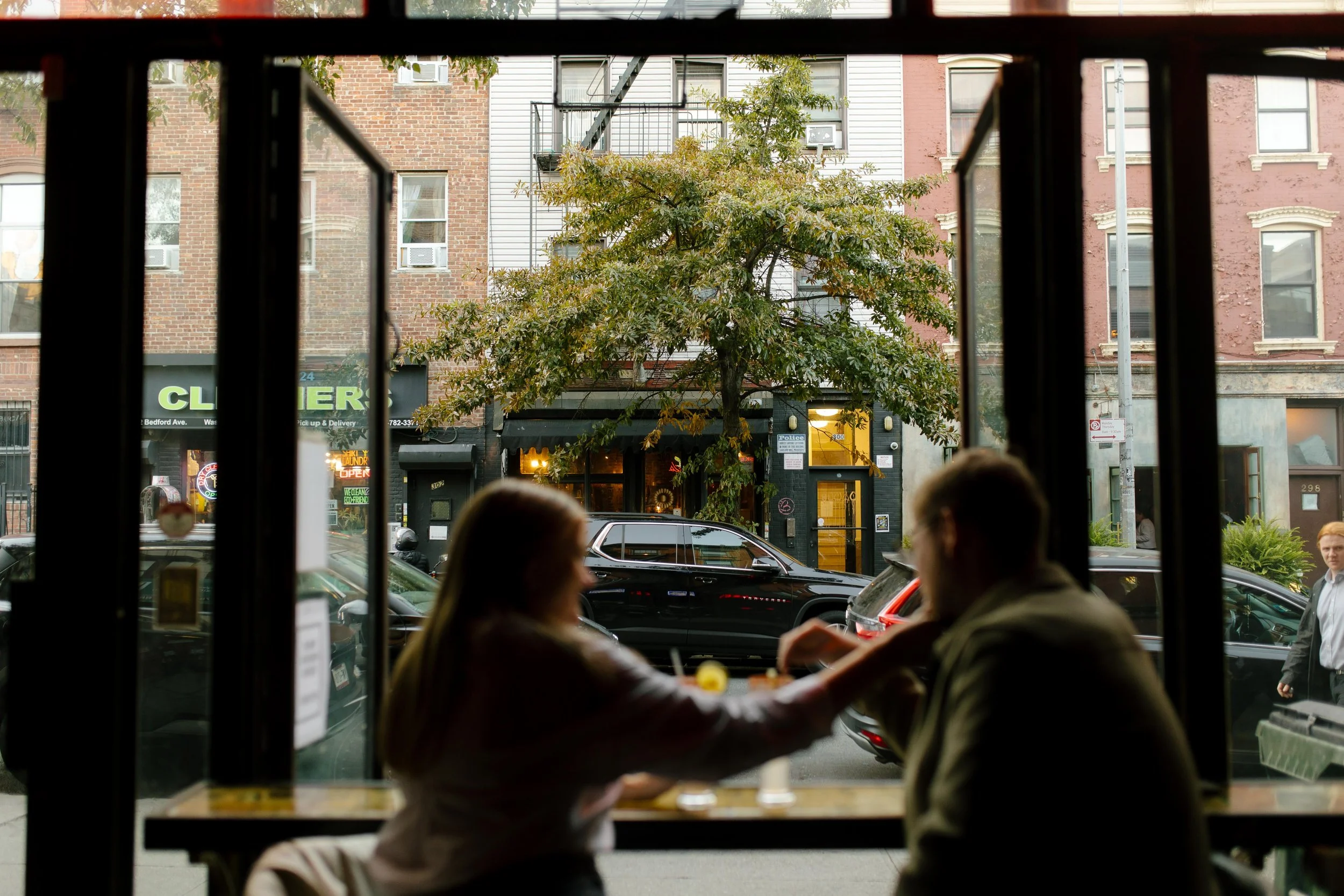 Couple reaching for each other across a window bar seat, Brooklyn street life visible outside.