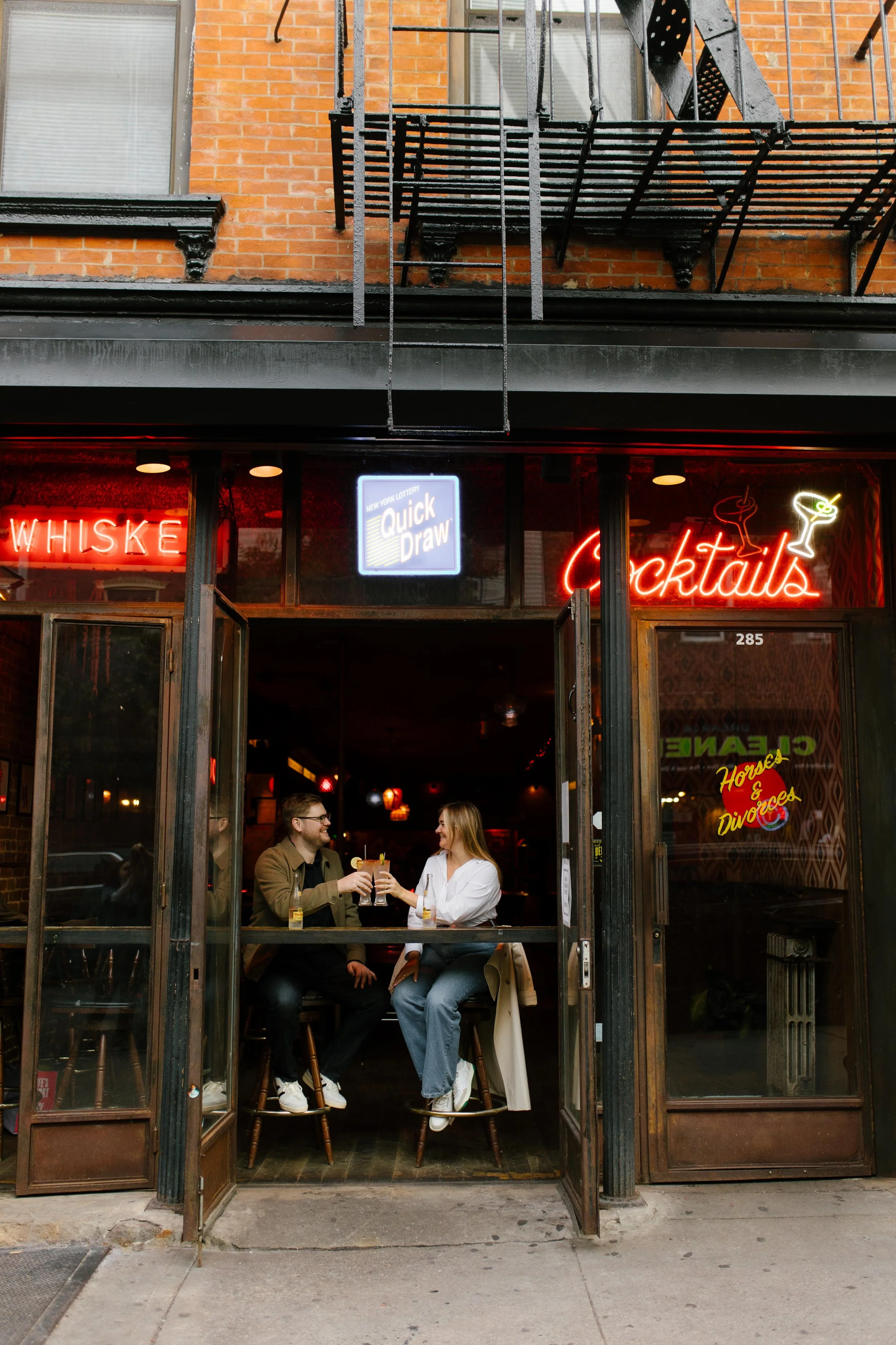 Couple toasting drinks inside a Brooklyn cocktail bar with glowing neon signs and open street doors.