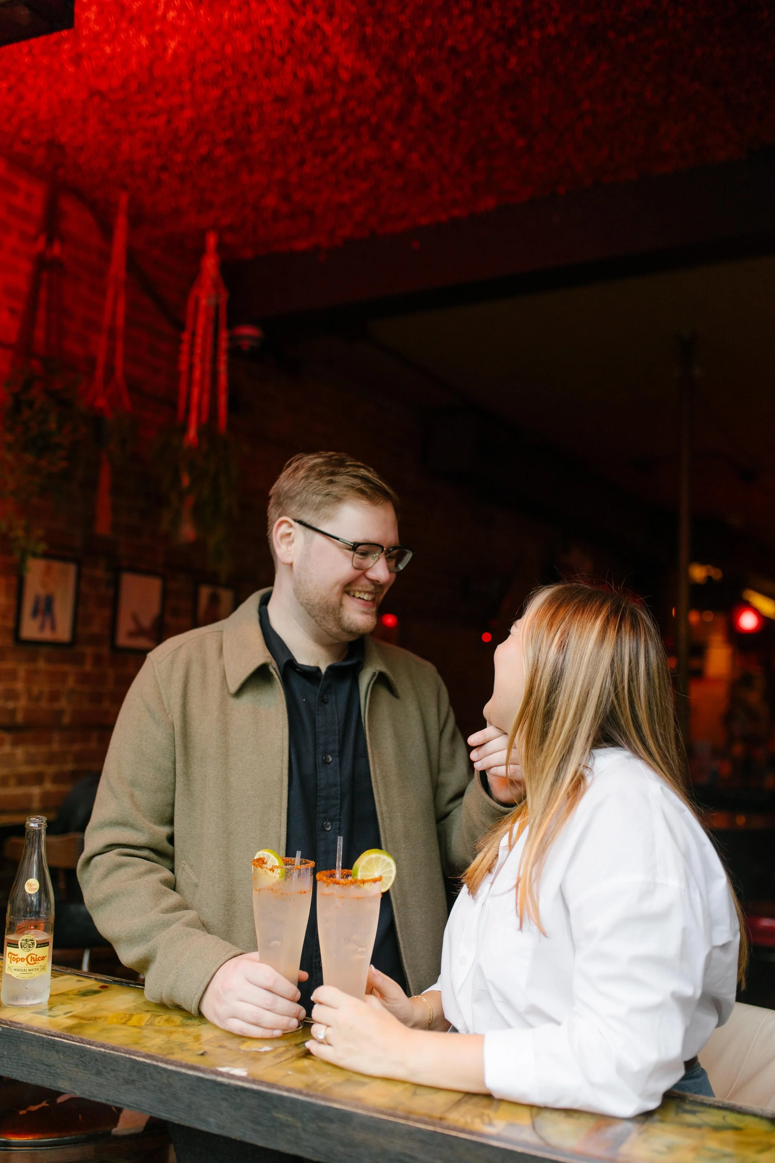 Playful nyc engagement photos inside a Brooklyn bar, couple holding cocktails beneath red neon lights.