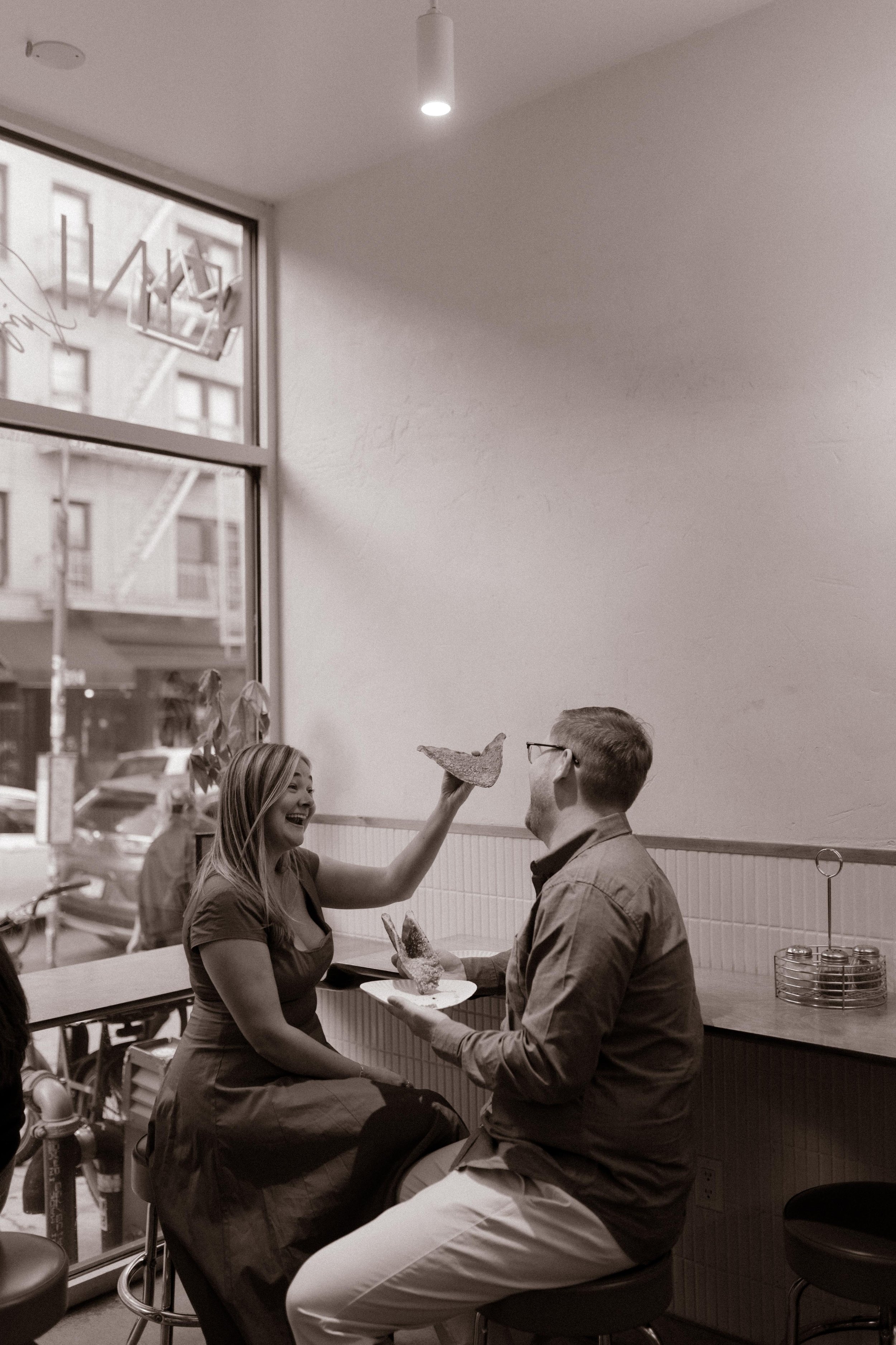 Black-and-white nyc engagement photos of couple laughing inside a Brooklyn pizza shop by the window.