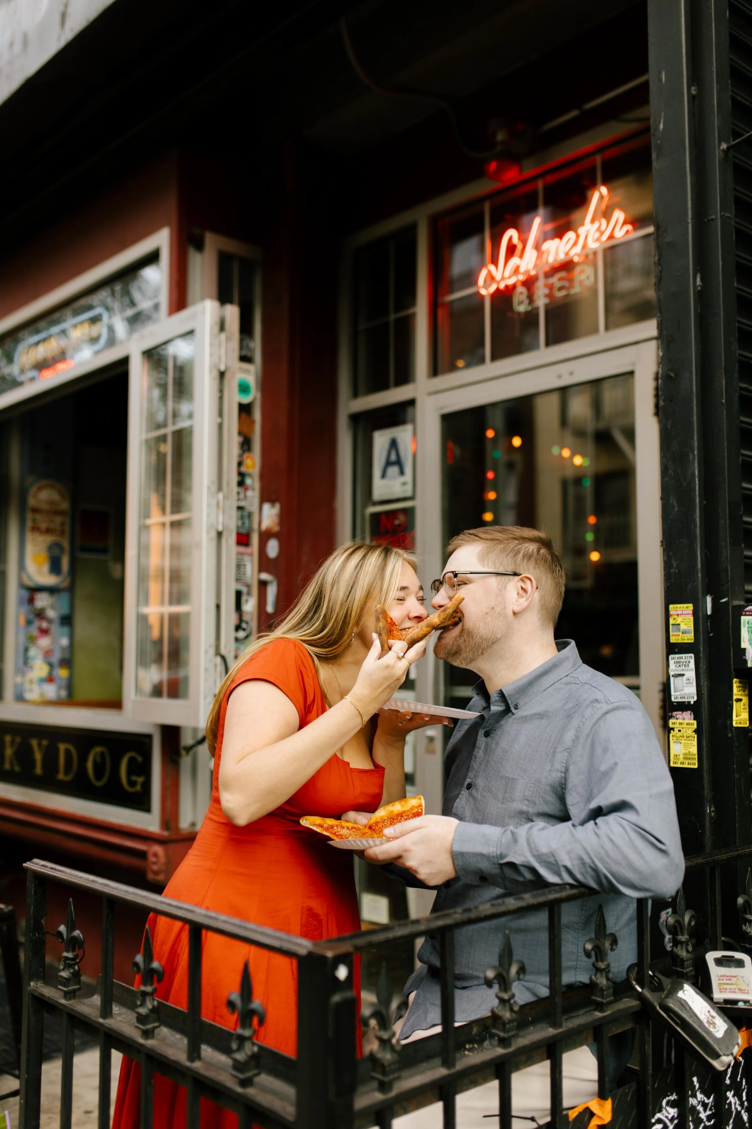 Playful nyc engagement photos of couple sharing pizza outside a Brooklyn bar beneath neon lights.