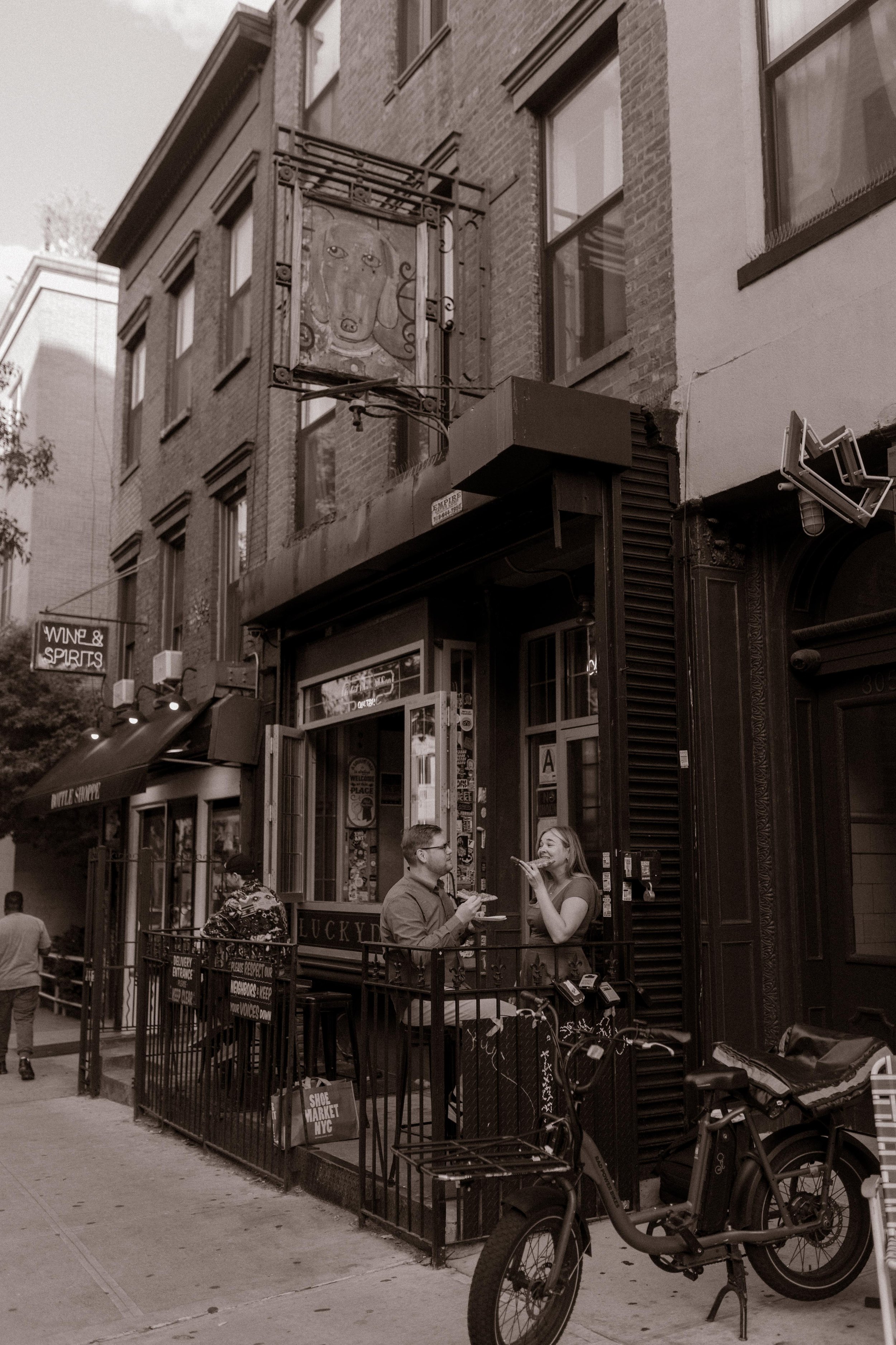Black-and-white street scene of couple eating pizza outside a Brooklyn bar during engagement session.