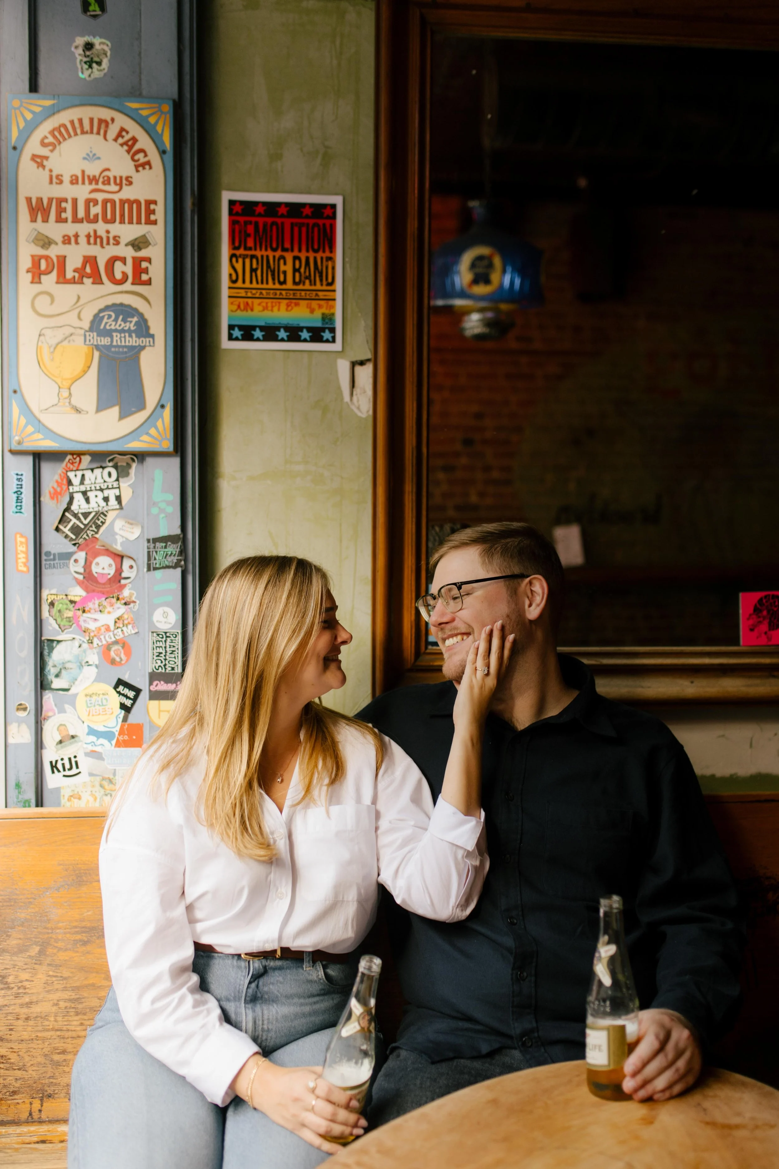Cozy nyc engagement photos inside a Brooklyn bar, couple smiling at each other over bottled beers.