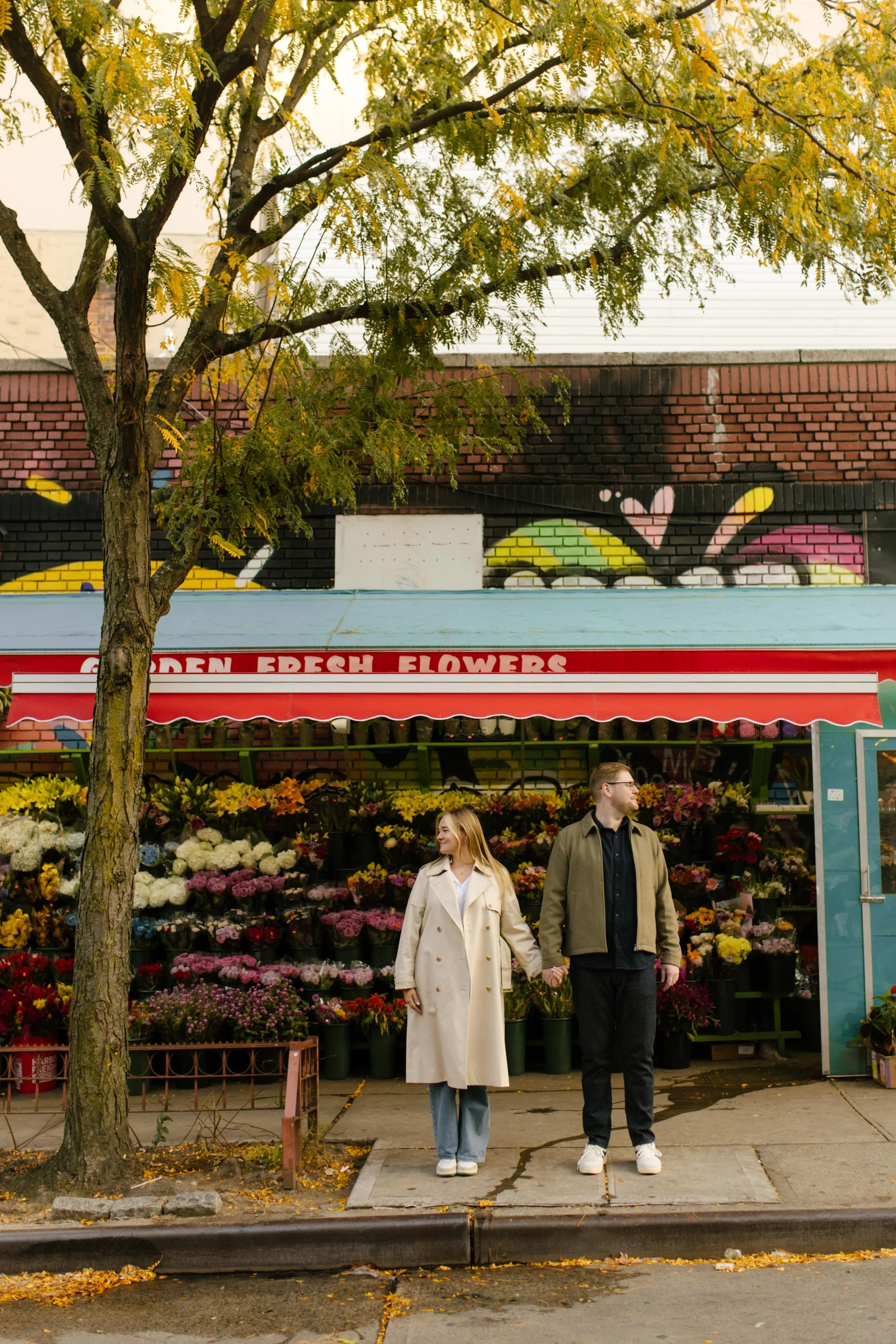 Couple standing hand-in-hand in front of vibrant Brooklyn flower shop during fall engagement session.