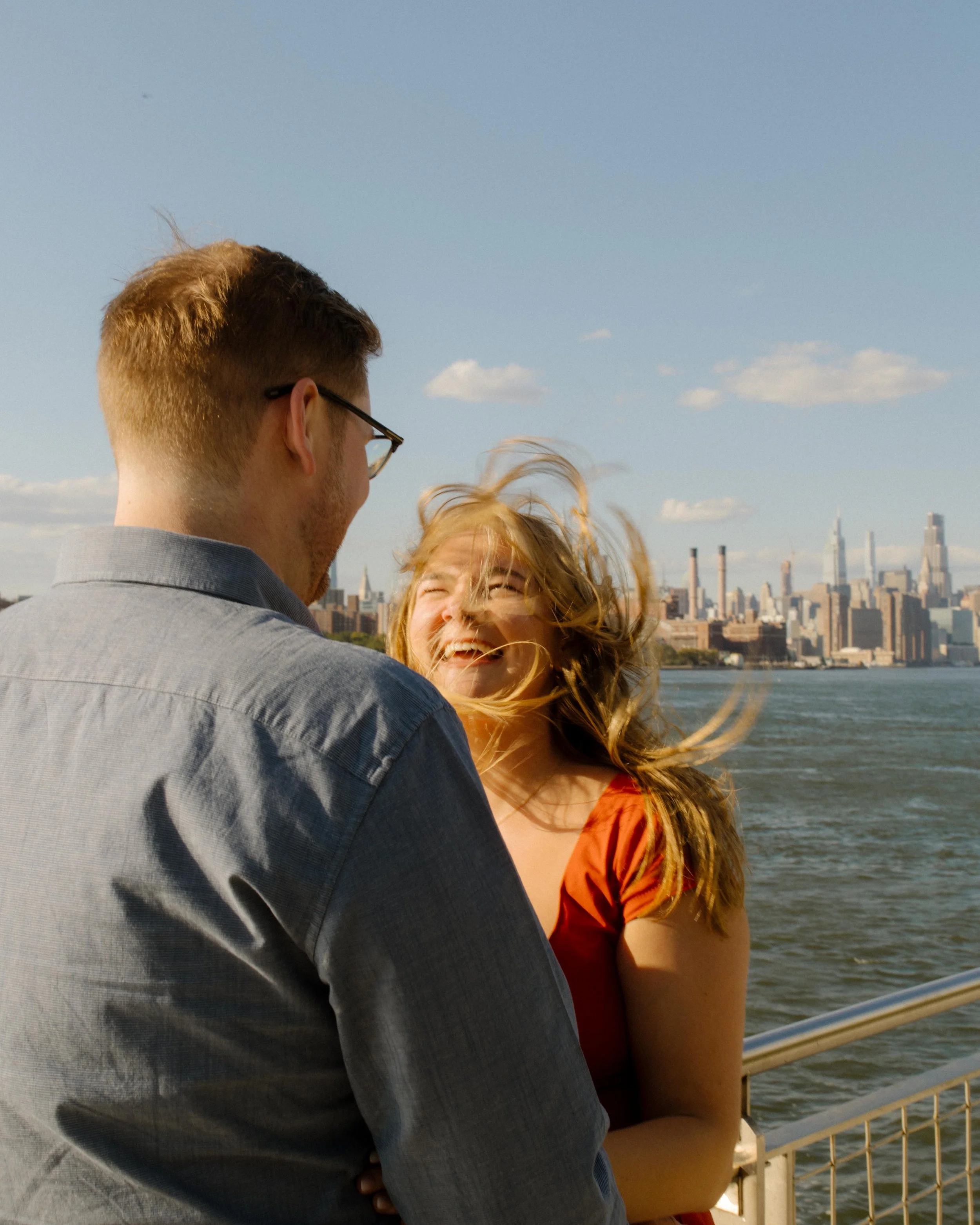 Wind-swept portrait of couple laughing by the water with the Manhattan skyline in the background.