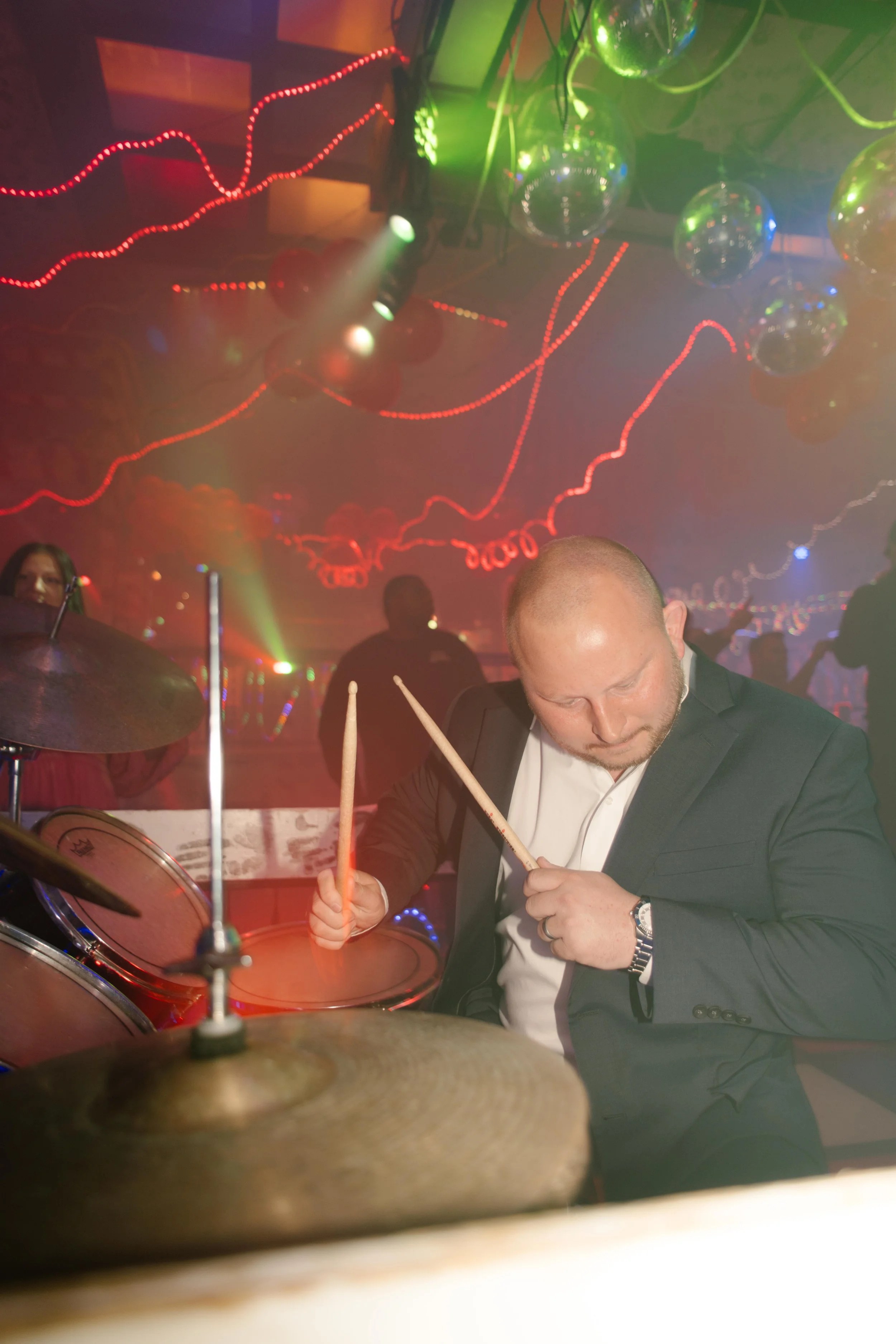The groom plays the drums in his suit under colorful club lights, sweat and joy mixing as the dance floor cheers him on during the wedding after party.