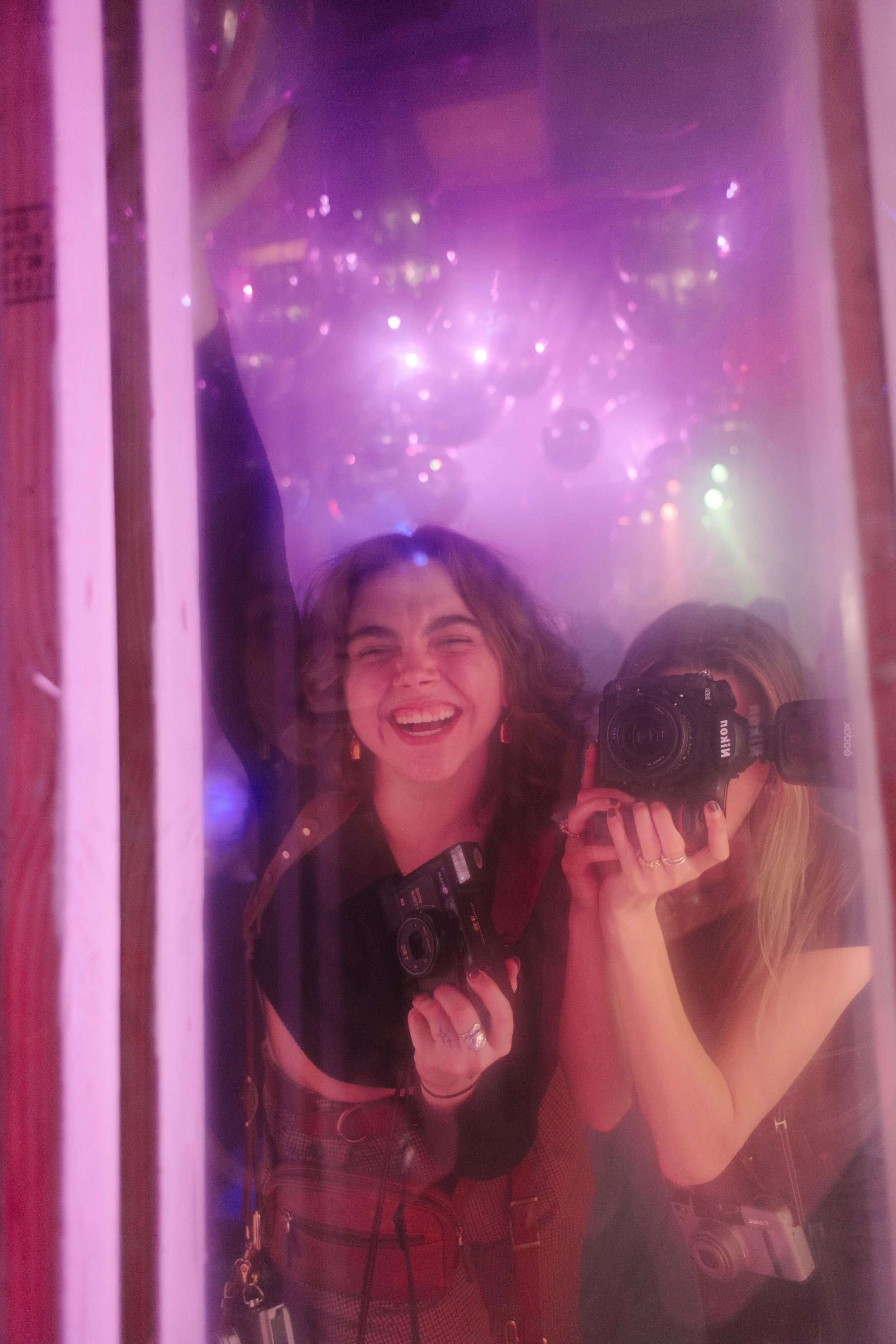 Two photographers laugh inside a mirrored booth filled with purple haze and disco balls, cameras in hand as they document the wild energy of wedding after parties.