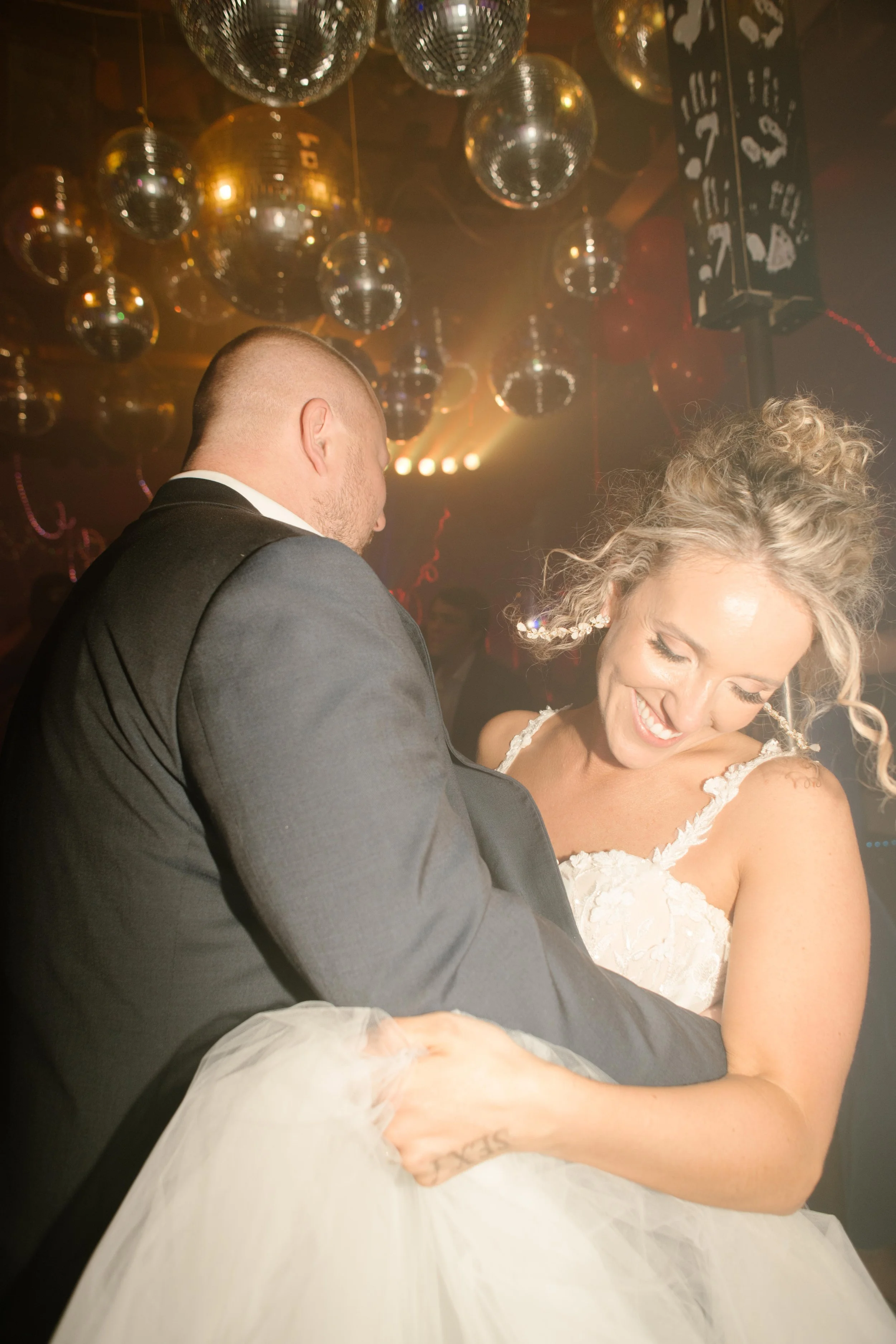 The groom pulls the bride close beneath a ceiling of disco balls as she smiles down at her tulle skirt, caught in a soft, golden haze on the dance floor.