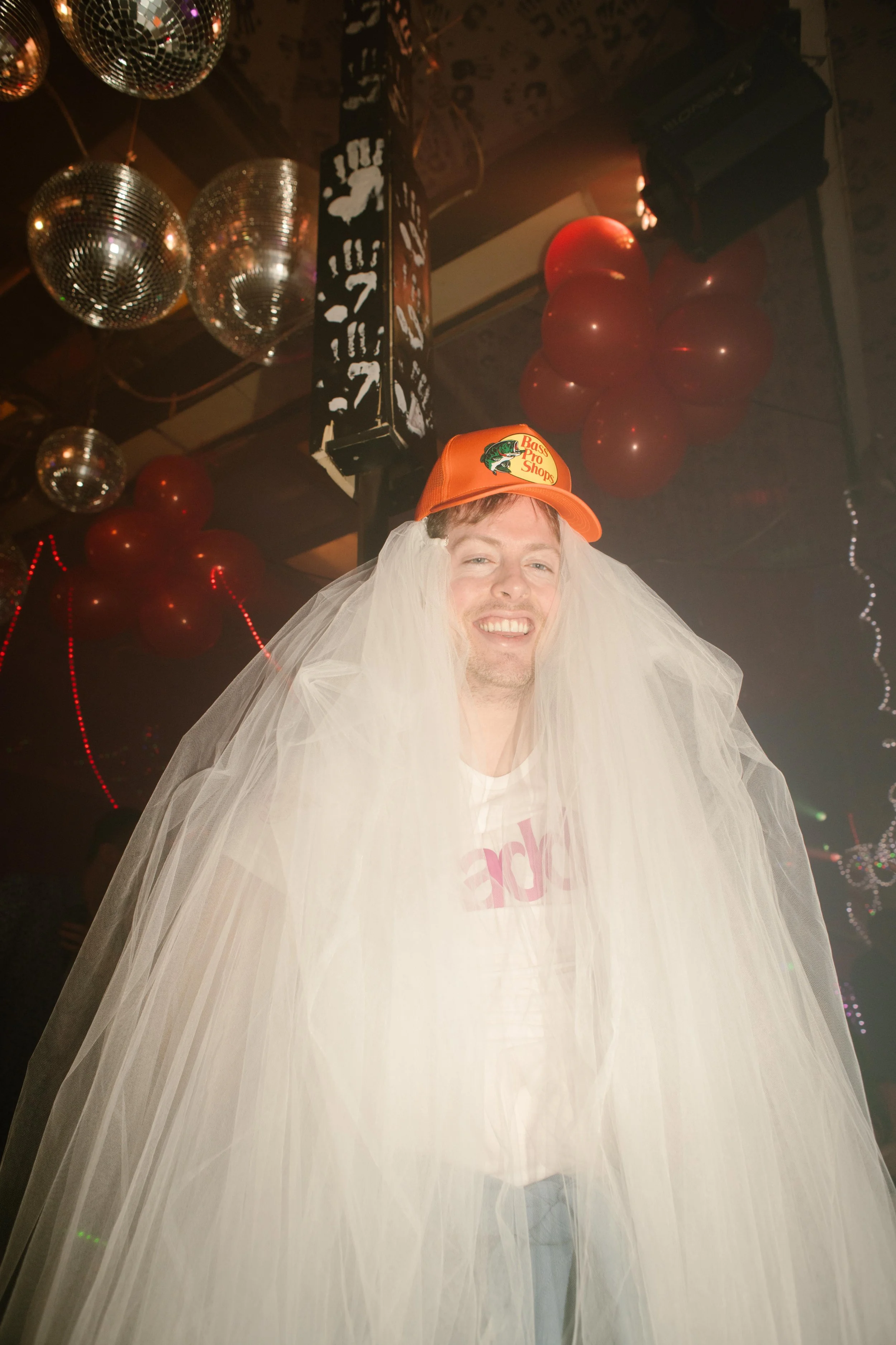 A smiling guest wears a baseball cap and a long bridal veil under disco balls and red balloons, embodying the playful spirit of wedding after parties.