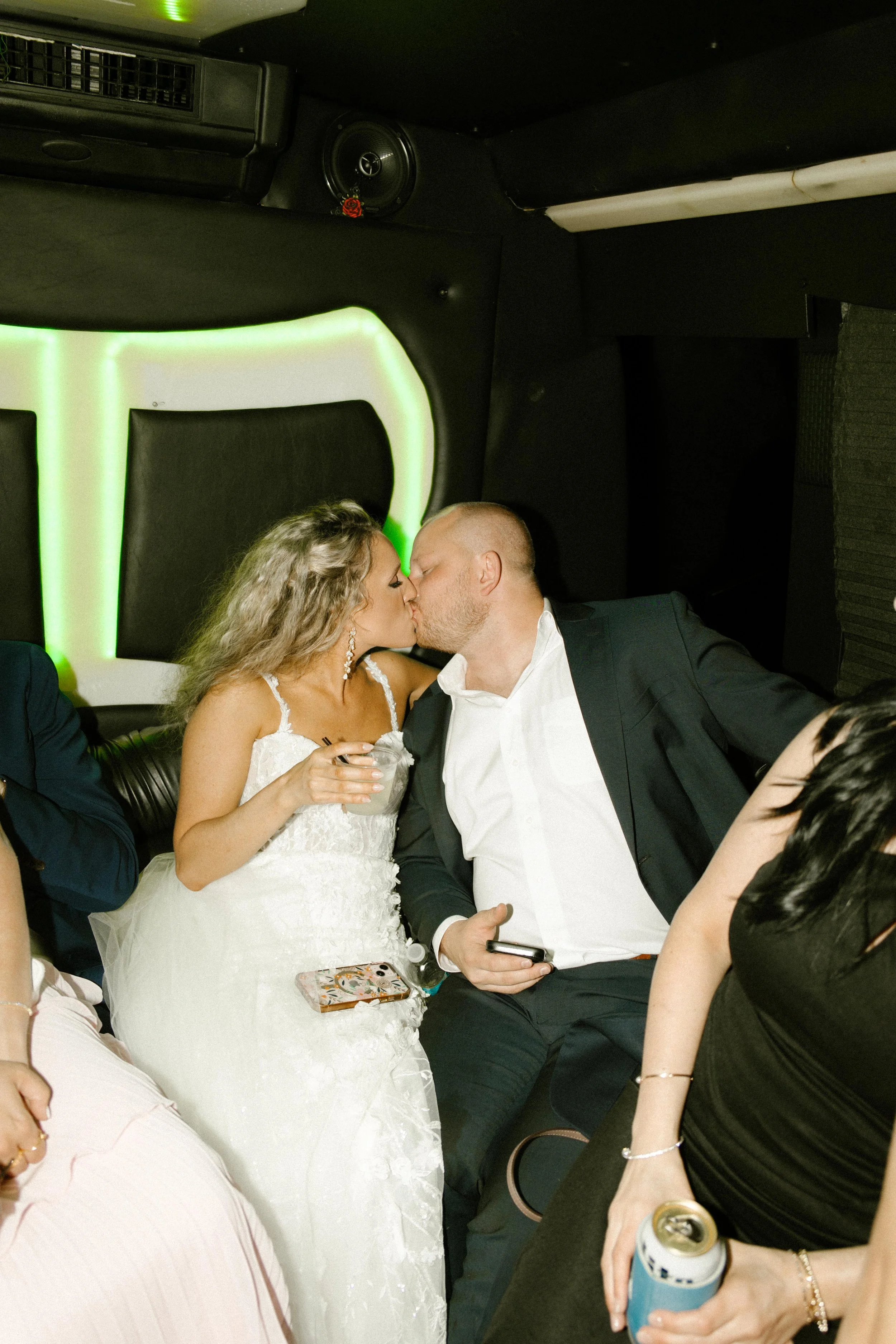 The bride and groom kiss in the back of a limo, her hand holding a cocktail while neon green lights glow behind them during the wedding after party.