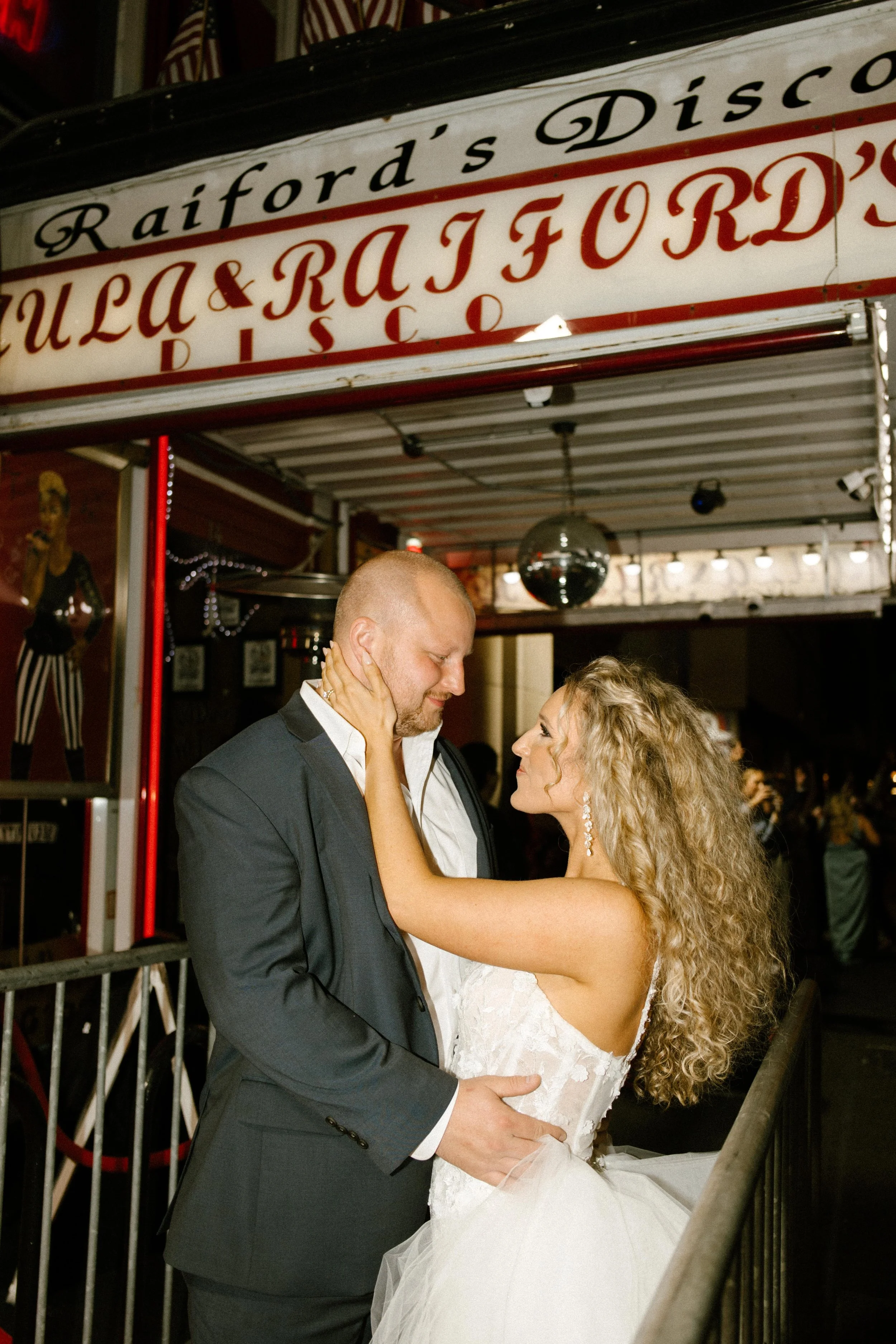 The bride and groom embrace beneath a vintage disco sign reading “Raiford’s Disco,” sharing a quiet moment on the balcony before heading back into the party.