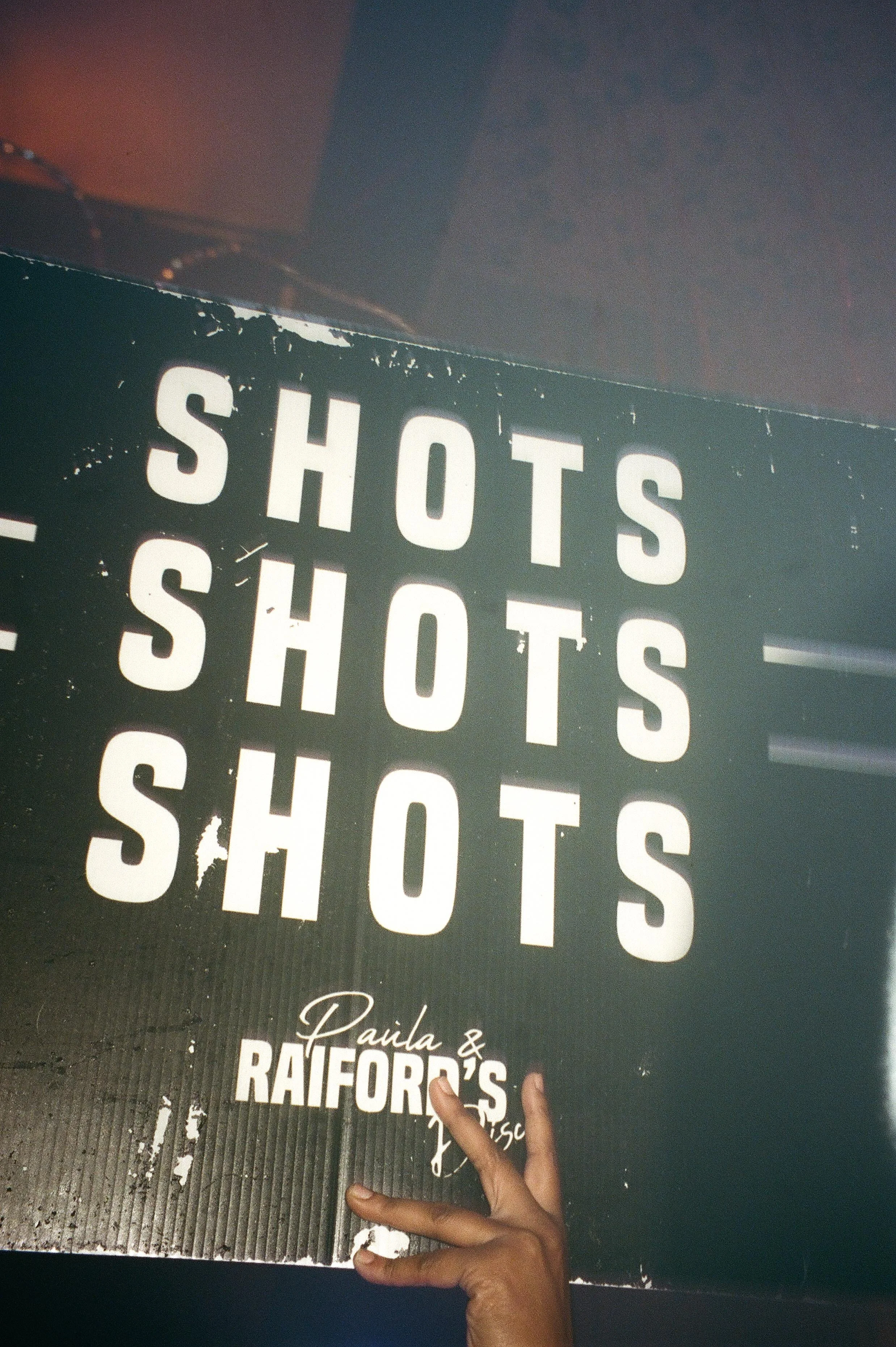 A worn black sign reading “SHOTS SHOTS SHOTS” with “Paula &amp; Raiford’s Disco” printed below, held up under warm, hazy club lighting during wedding after parties.