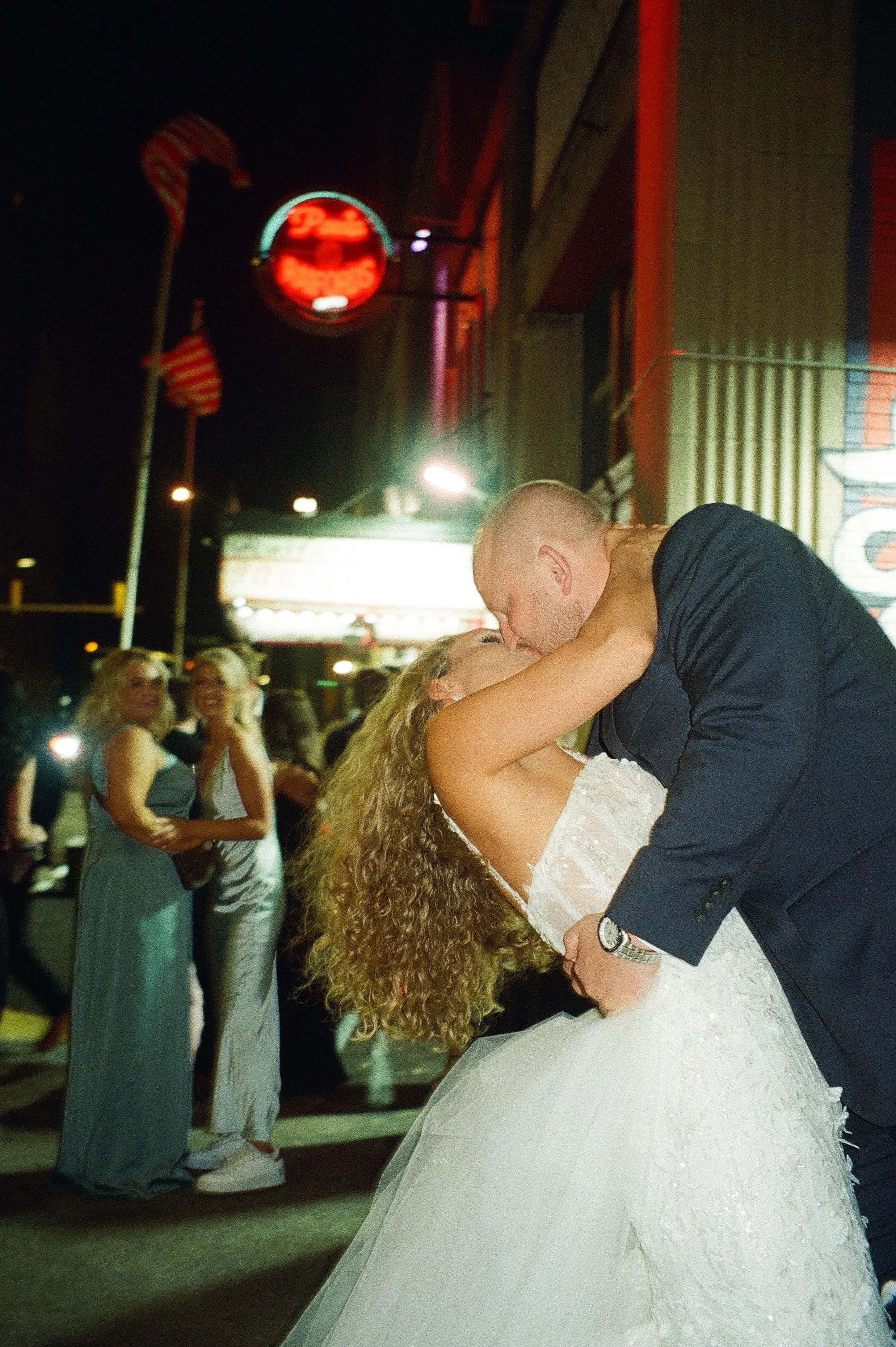Outside beneath a neon bar sign, the groom dips the bride into a kiss on the sidewalk while bridesmaids watch and smile during the wedding after party.