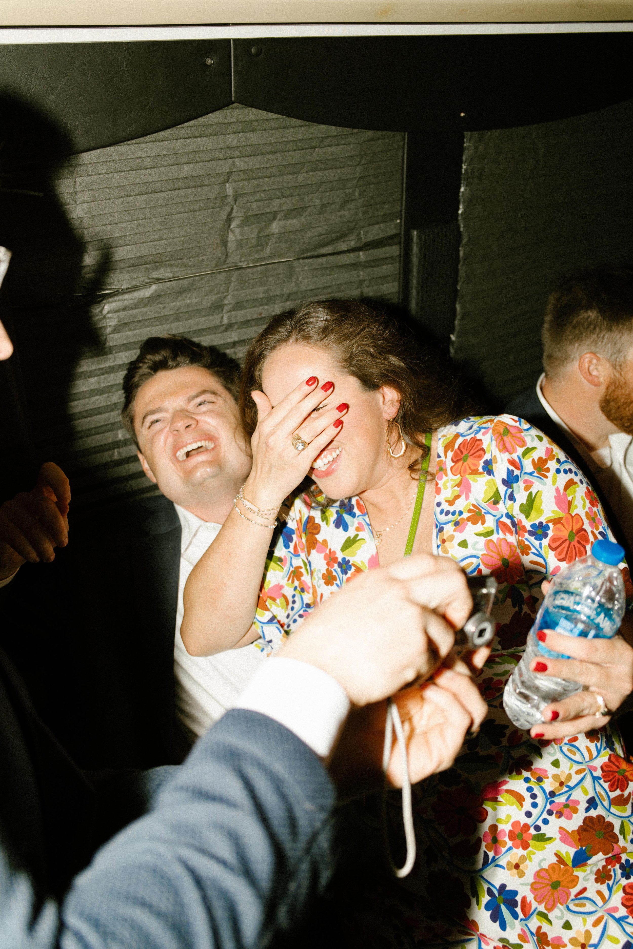 Guests packed into a photo booth laugh wildly, one woman covering her face with bright red nails while holding a water bottle, capturing the chaotic joy of wedding after parties late into the night.