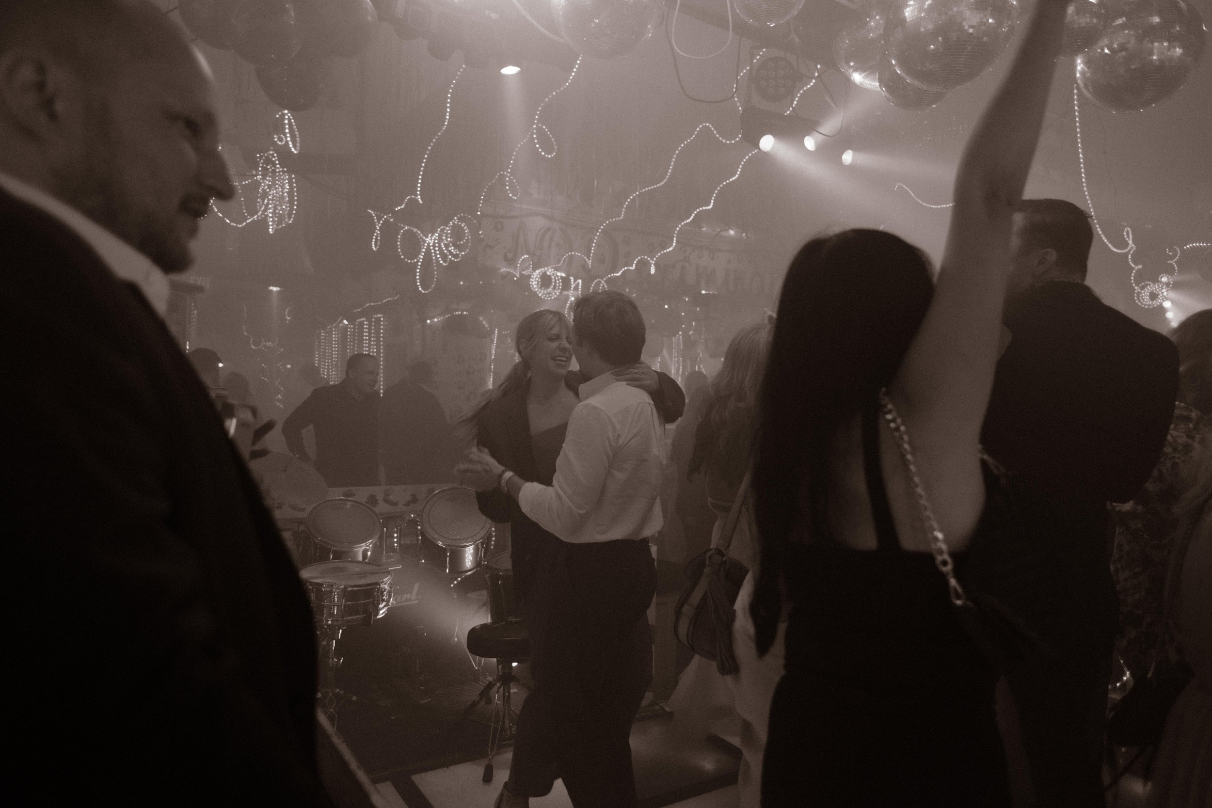On a crowded dance floor under disco balls and string lights, guests sway and embrace in the smoky glow of an epic wedding after party.