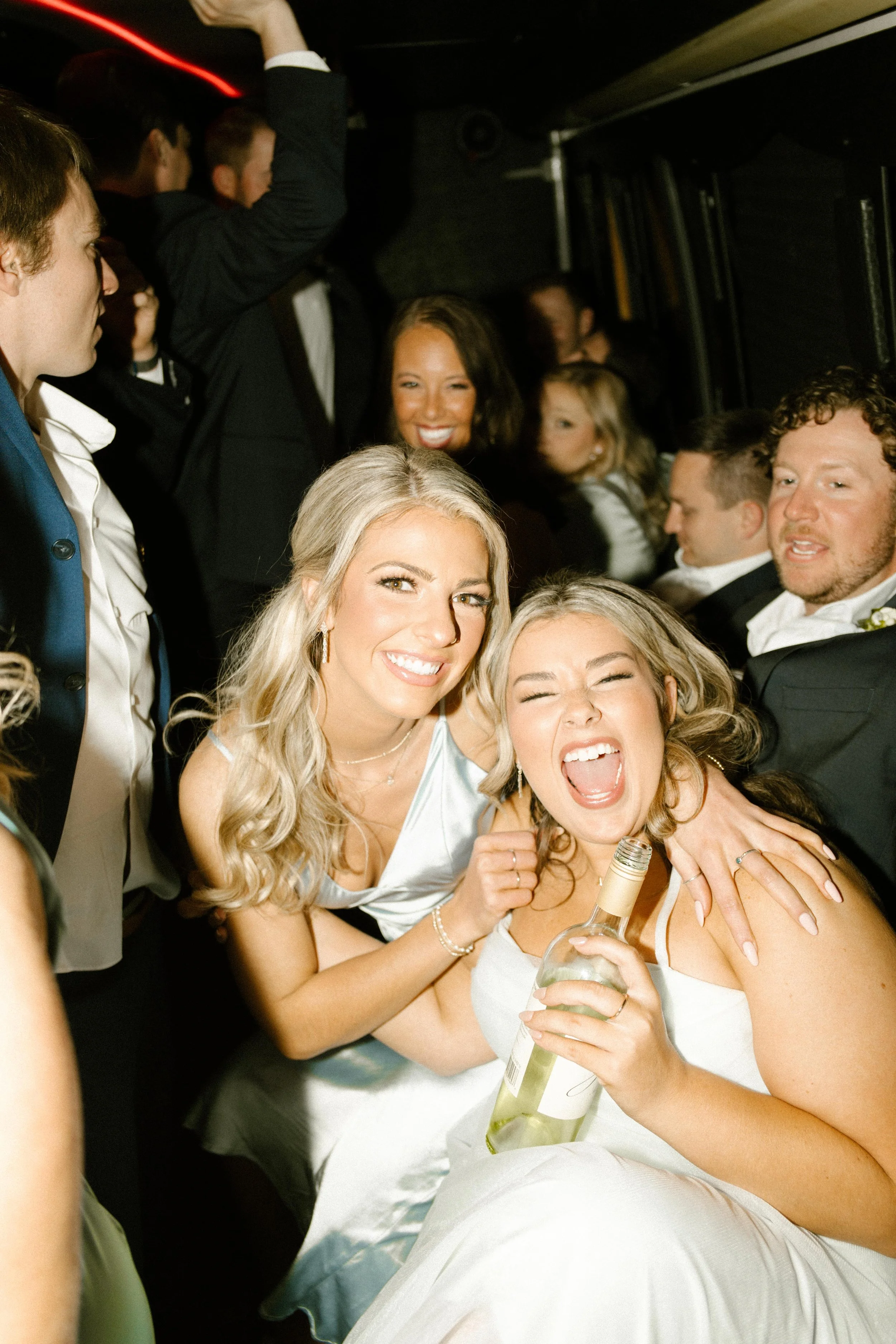 Two bridesmaids hugging and laughing with a wine bottle on a crowded dance floor during wedding after parties.