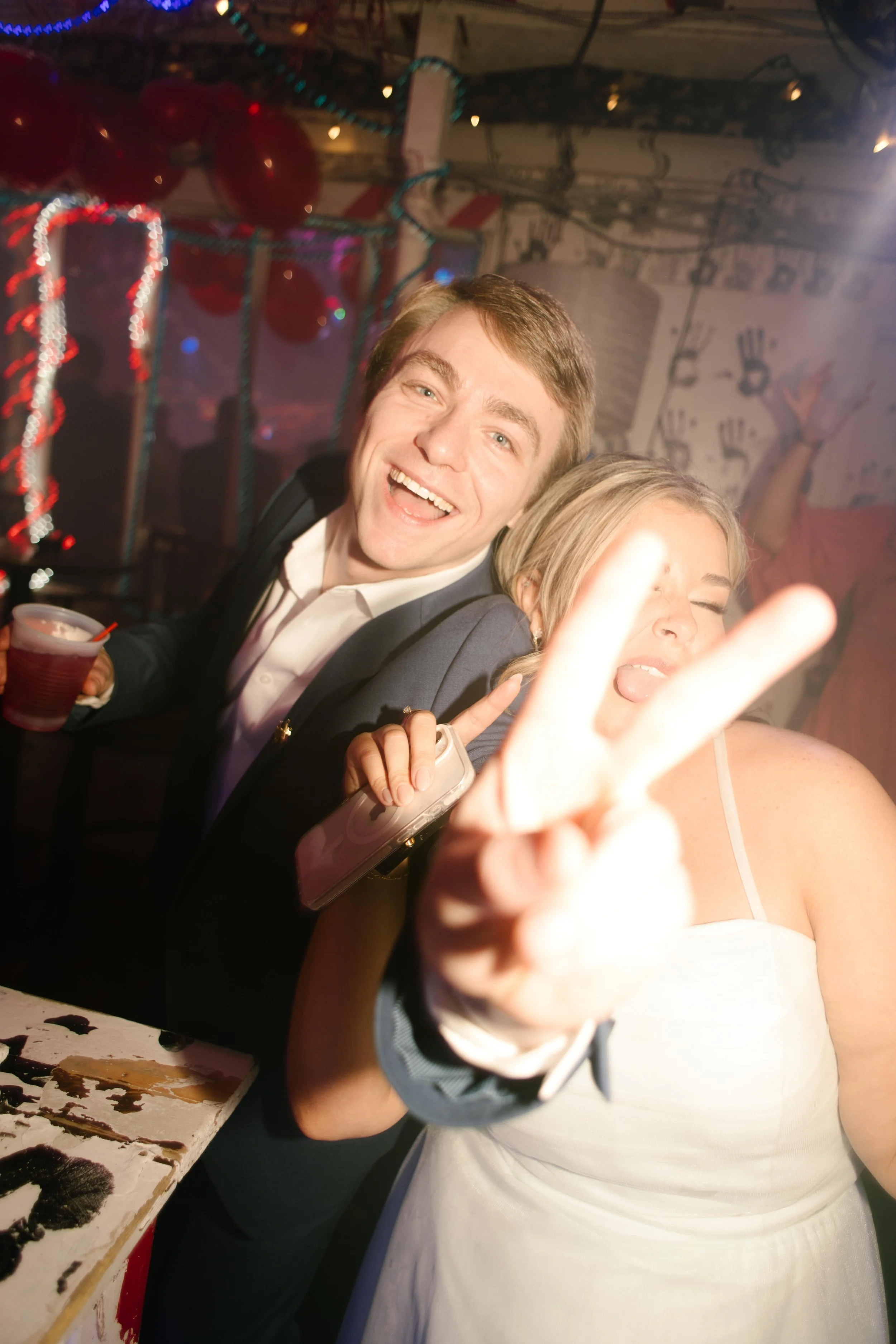 Two wedding guests smiling and posing with a peace sign while holding drinks on the dance floor.