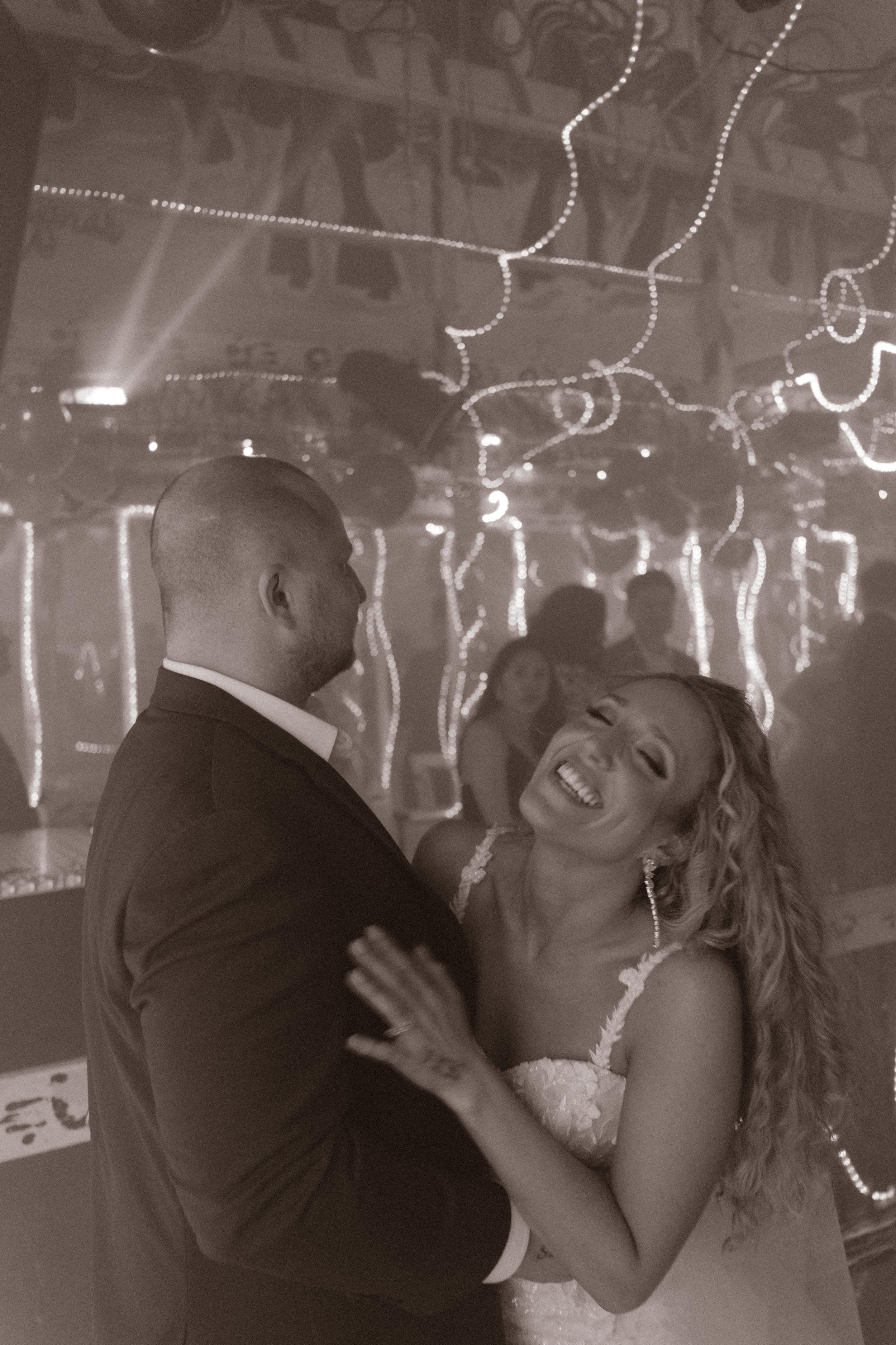 Bride laughing and dancing with the groom under glowing lights during wedding after parties.