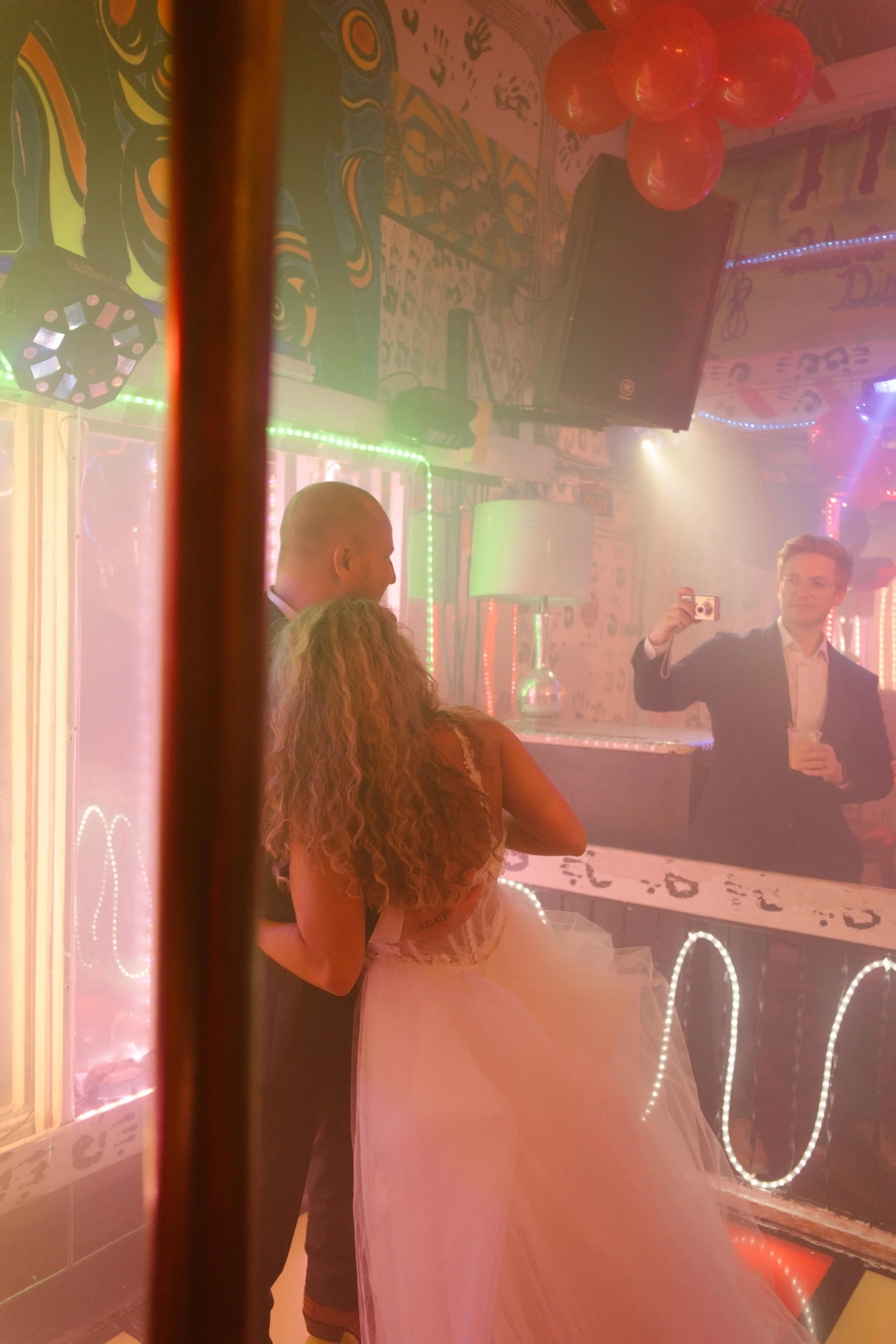 Friend taking a photo of the bride and groom dancing together inside a lively nightclub reception.