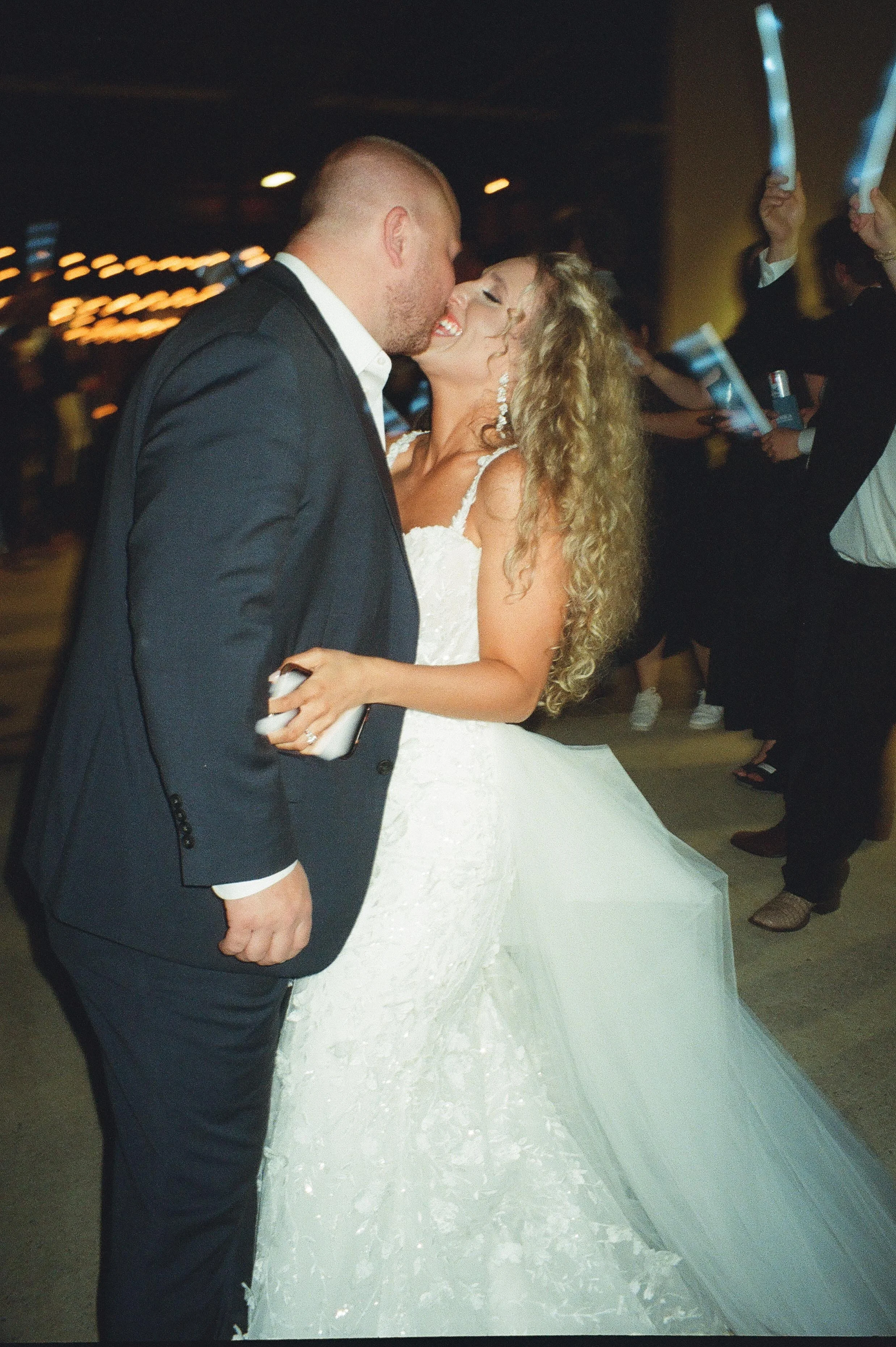 Newlyweds kissing while guests wave glow sticks during an energetic wedding after party exit.