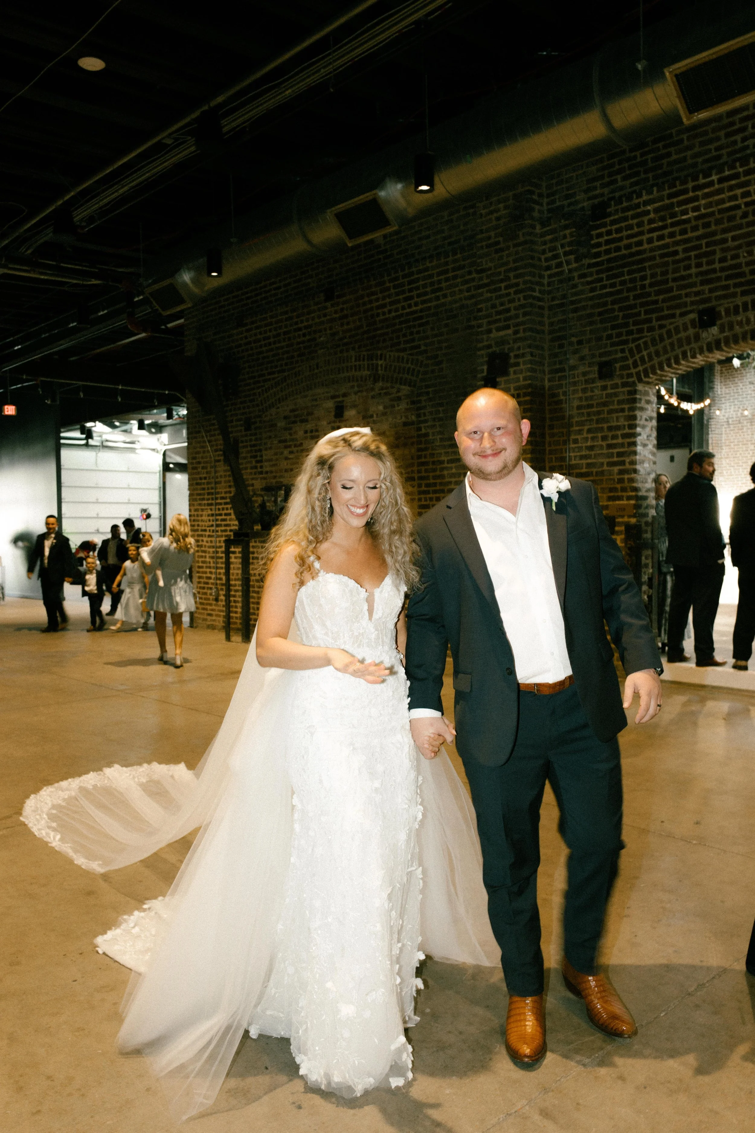 The bride and groom walk hand in hand through an industrial brick venue, her veil trailing behind them as they sneak away toward the wedding after party.