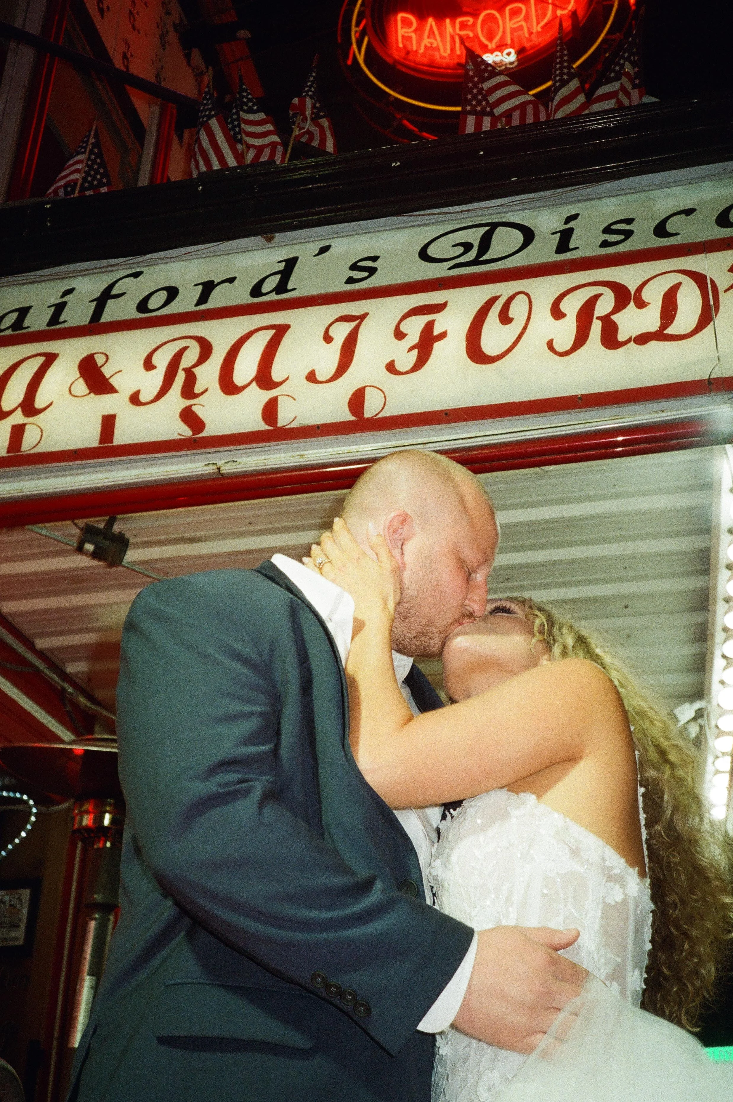 Beneath the glowing red Raiford’s sign, the groom dips the bride into a kiss outside the club, neon reflecting off their faces during wedding after parties.