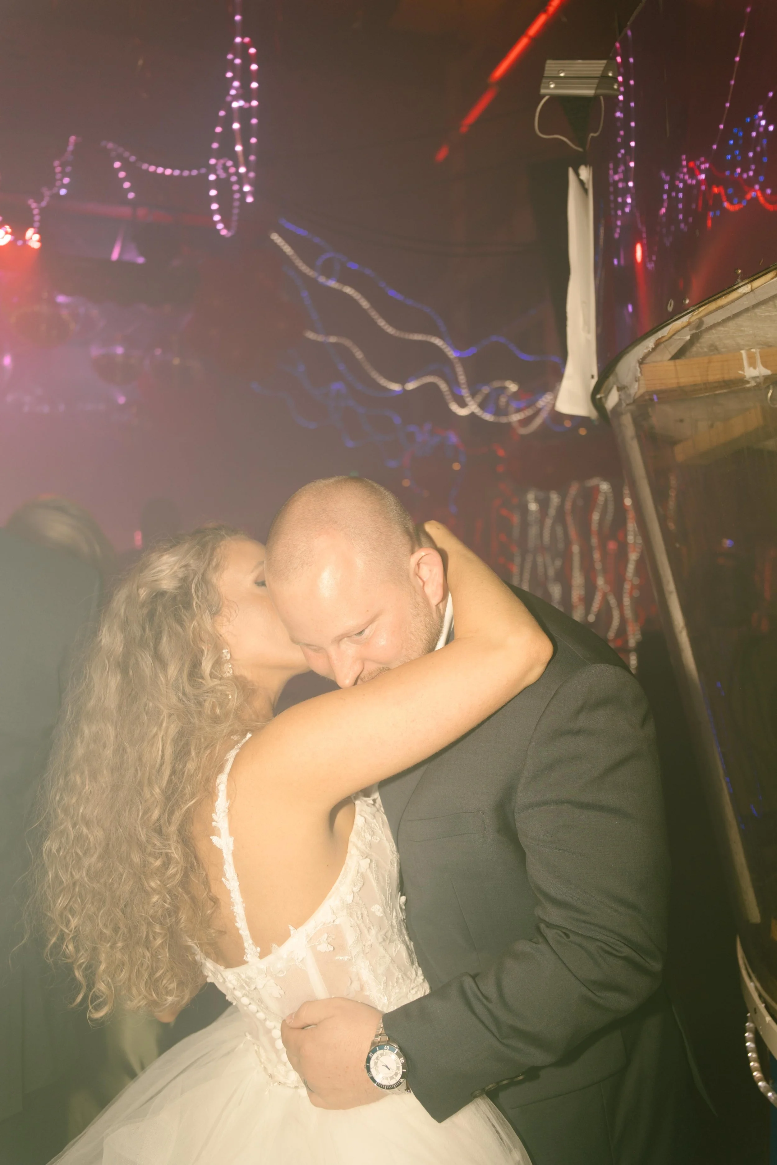 The bride wraps her arms around the groom’s neck and kisses his cheek in a hazy nightclub corner, disco lights glowing around them during the wedding after party.
