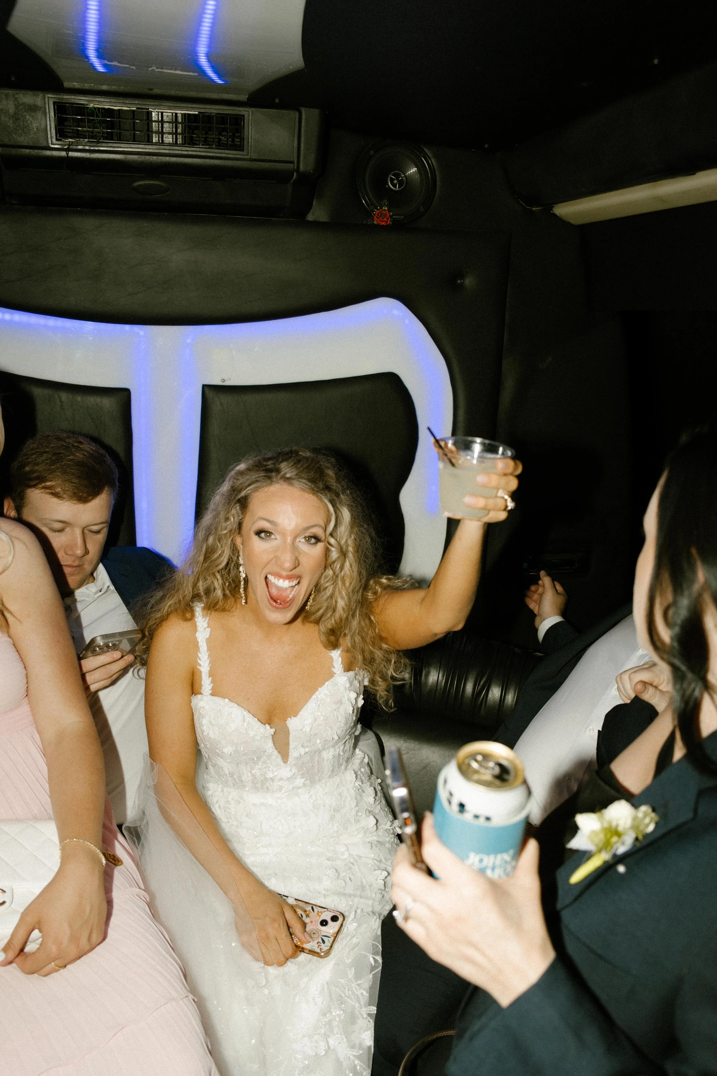 Bride raising a drink and cheering with friends inside a limo on the way to wedding after parties.
