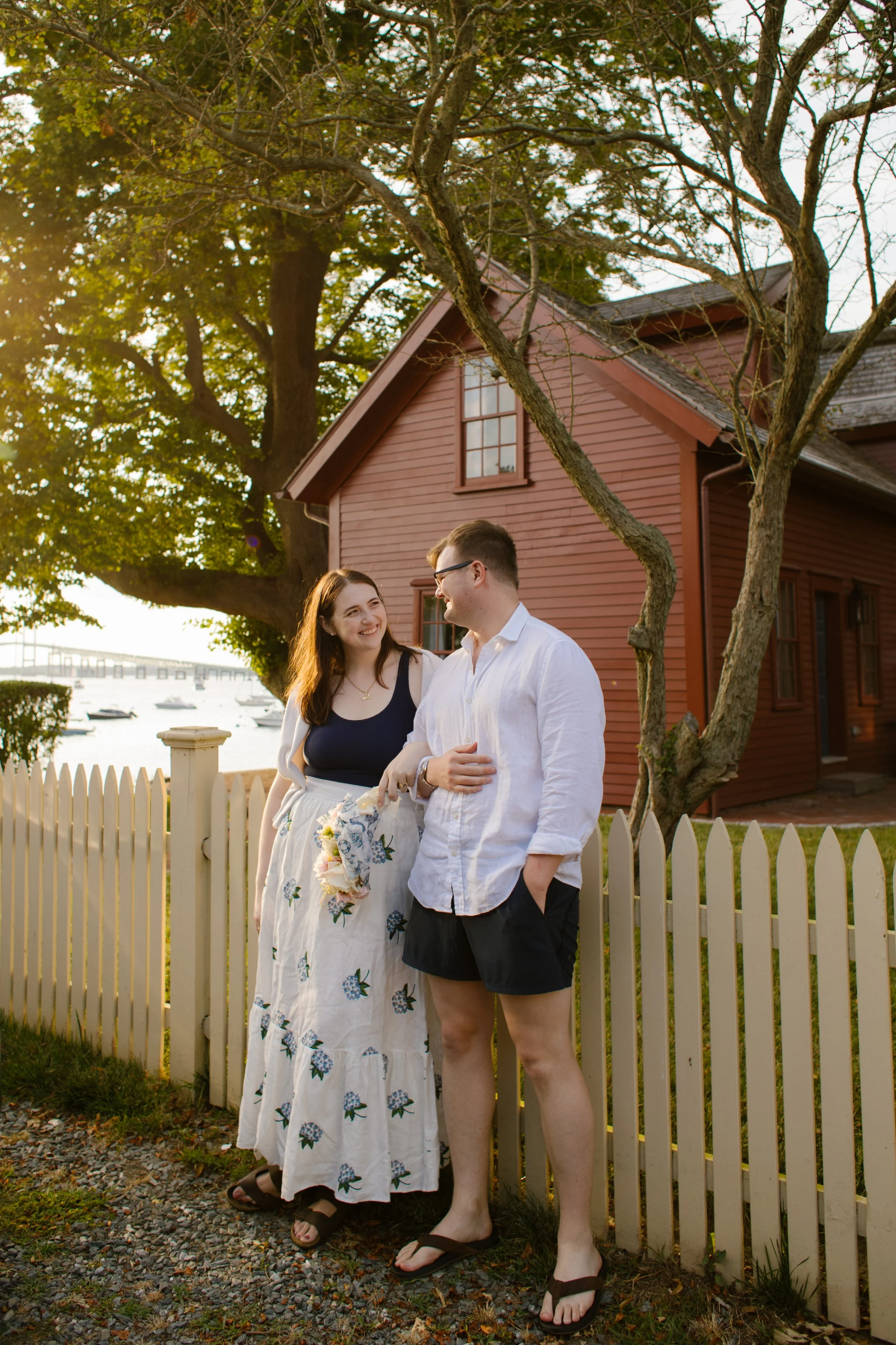 Couple standing by white picket fence and red cottage during Newport Rhode Island engagement photos