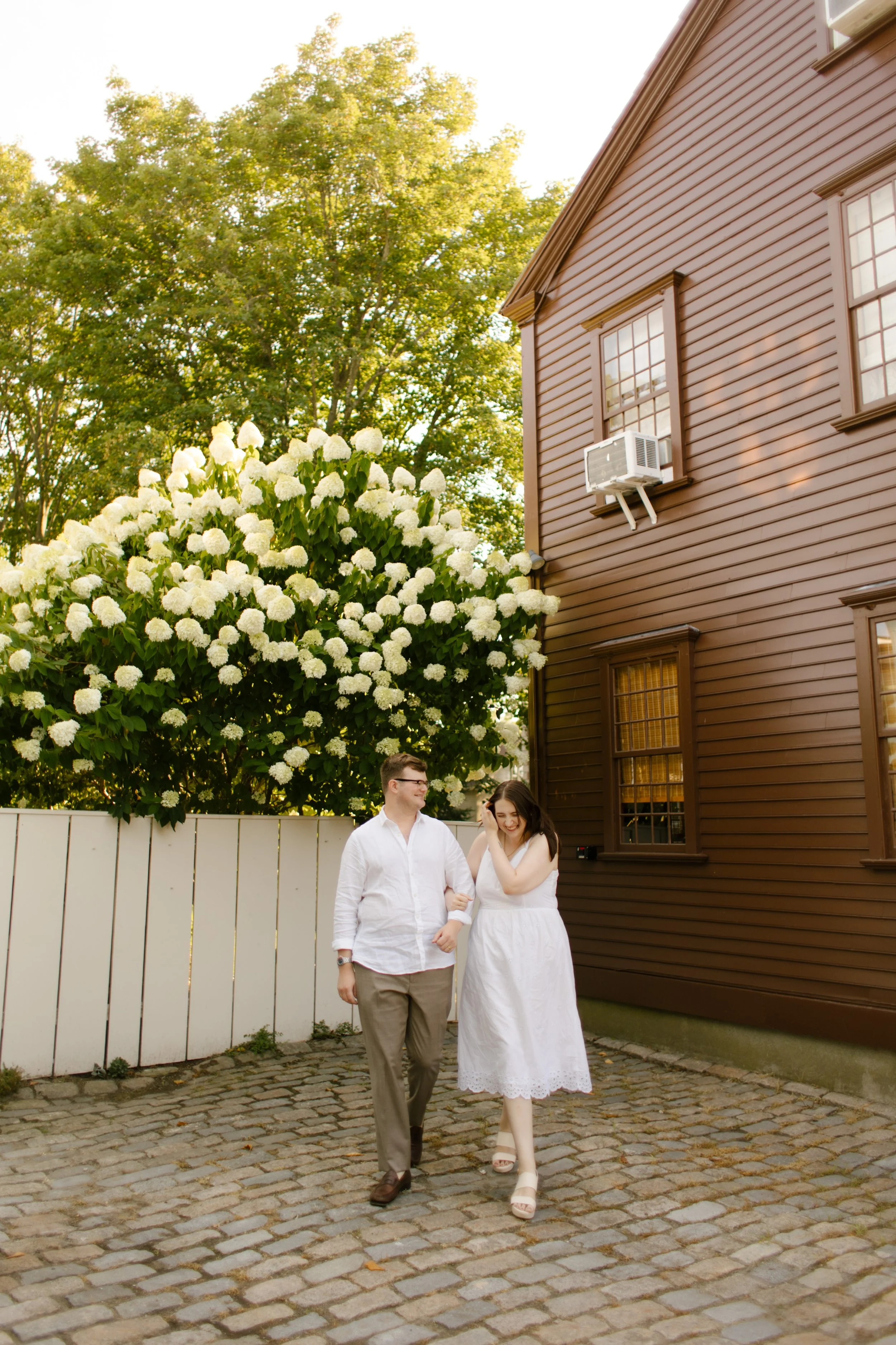 Couple walking past white summer blooms and historic brown Newport house