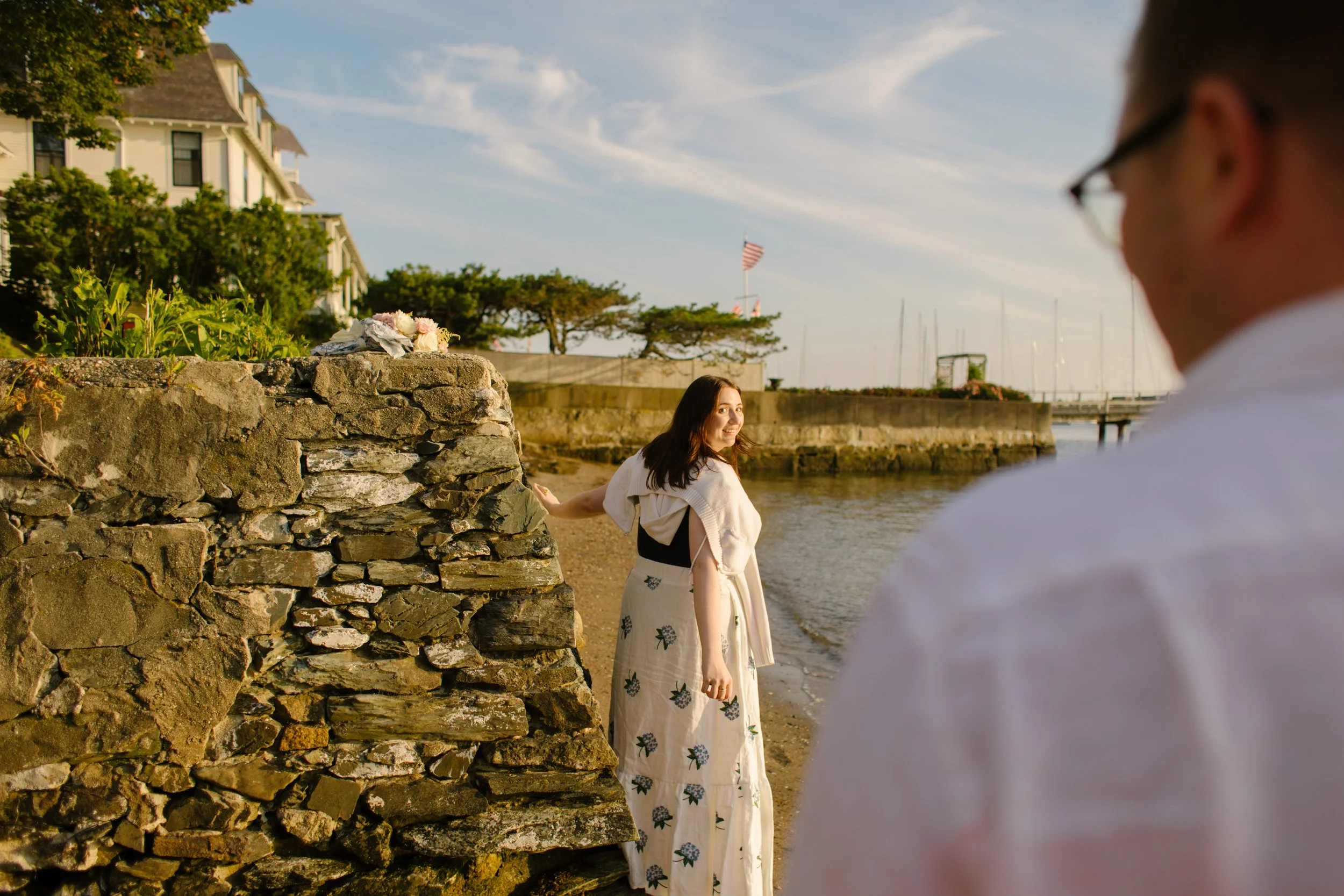 Bride-to-be standing by historic Newport harbor stone wall at golden hour