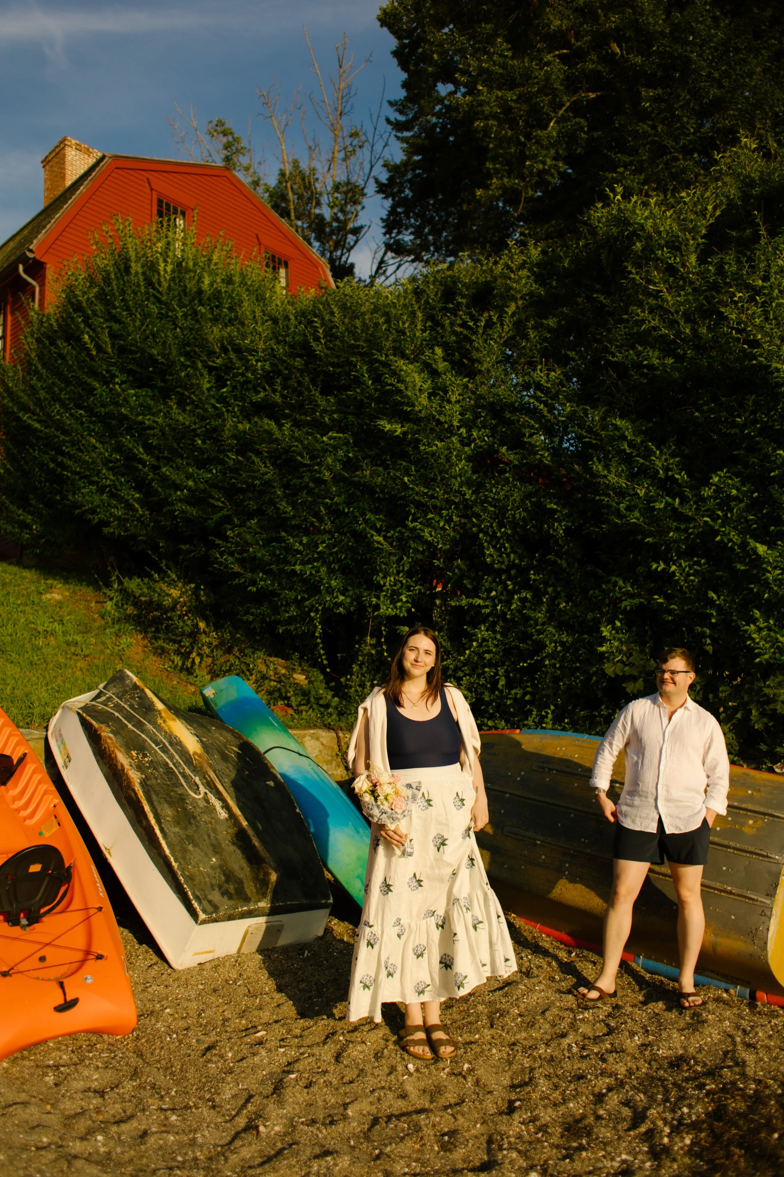 Couple standing on rocky shoreline with boats and red Newport house during Newport Rhode Island engagement photos