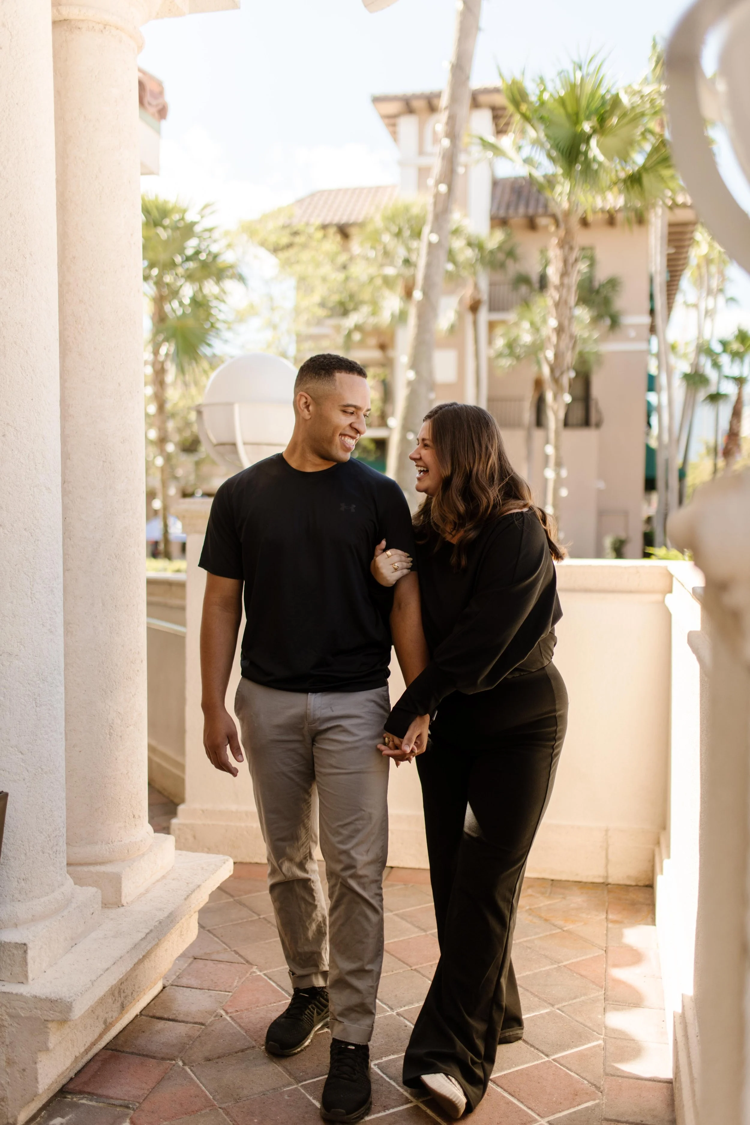 Couple walking hand in hand through a palm-lined walkway, laughing mid-step in warm afternoon light, a candid moment that feels like the in-between magic of how to plan a surprise proposal