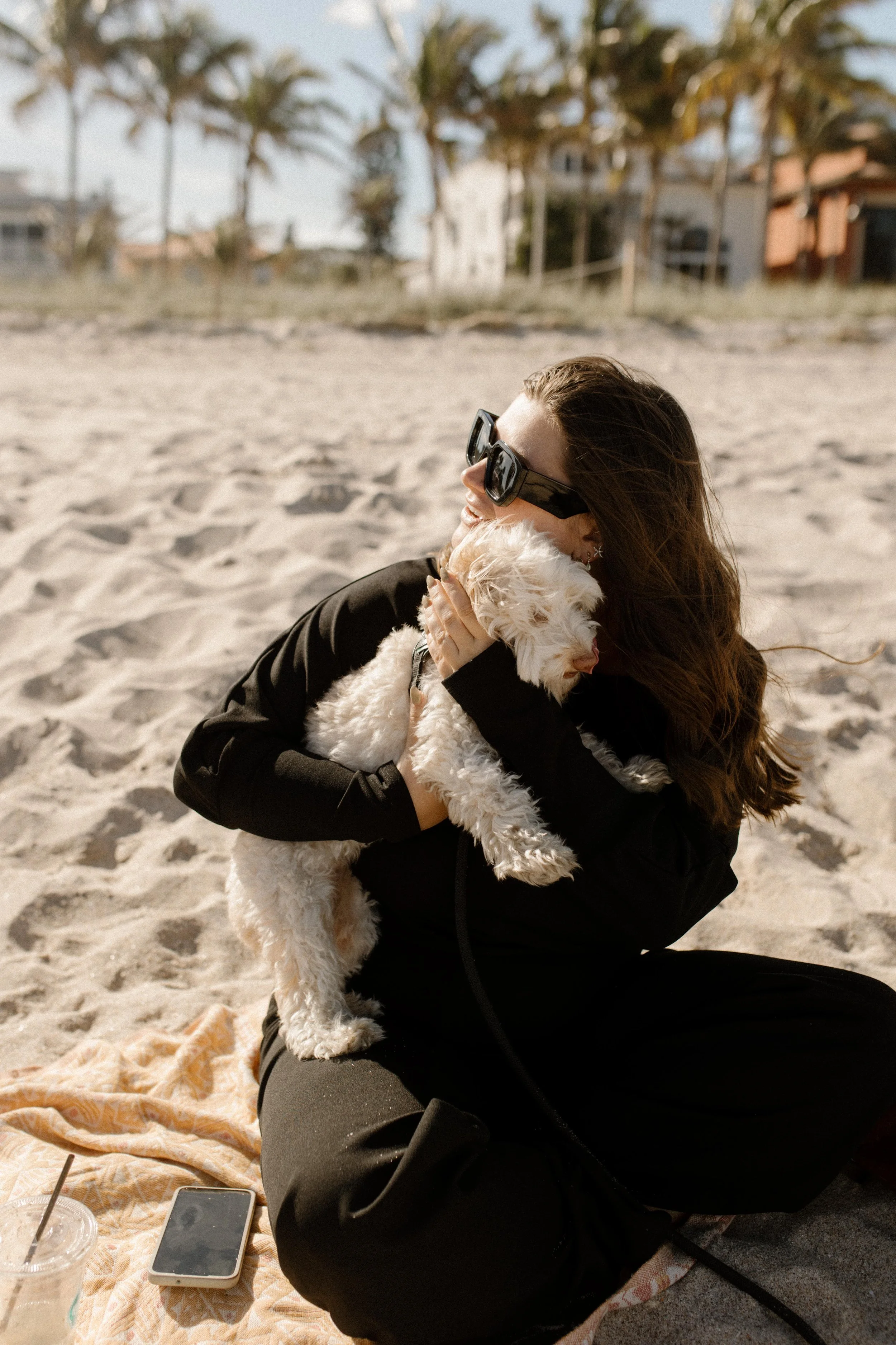 Woman sitting on the beach holding a small white dog, smiling in oversized sunglasses with wind in her hair and sunlight hitting the sand around her