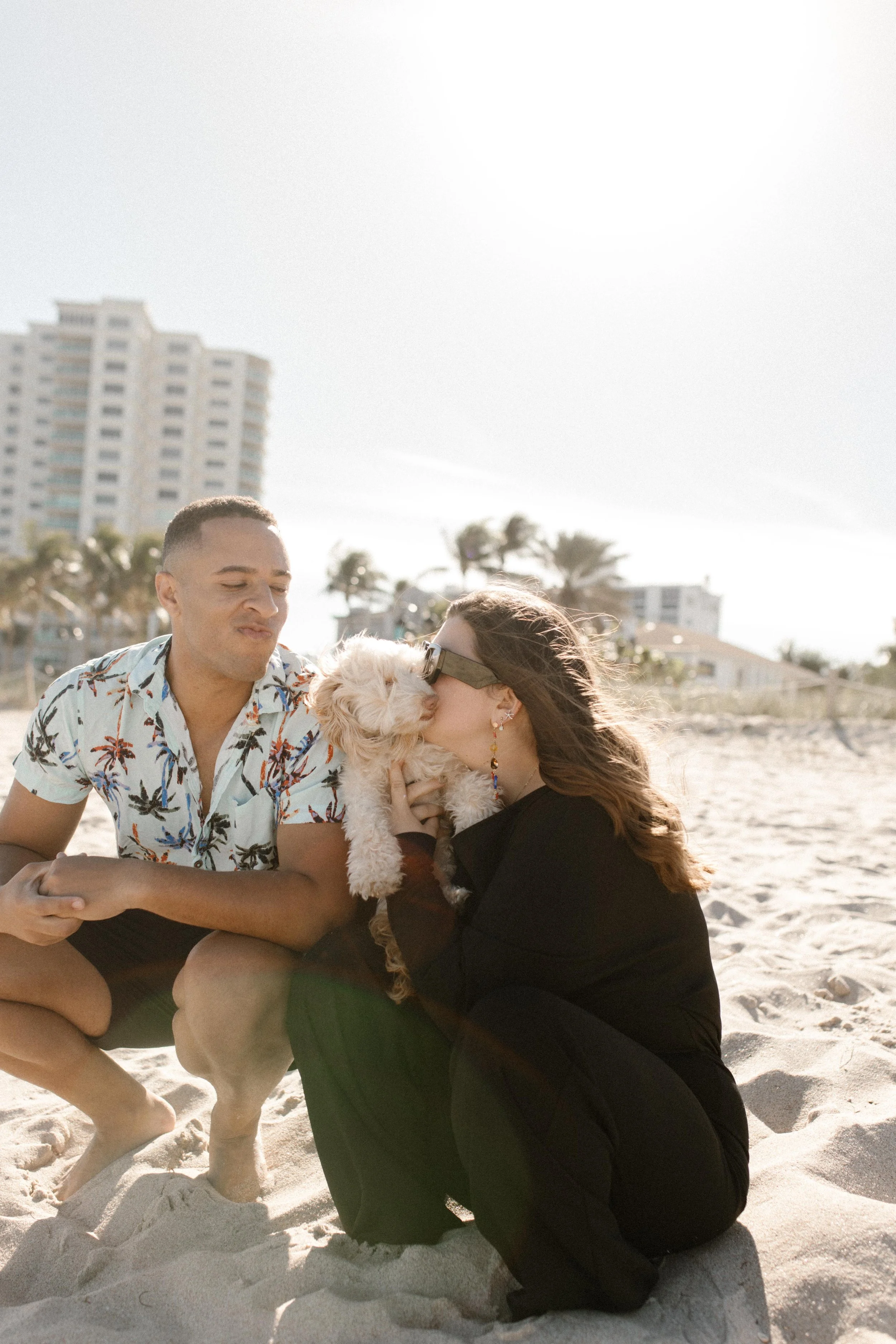 Woman kissing a fluffy white dog while sitting on the sand next to her partner like a carefree chapter in how to plan a surprise proposal